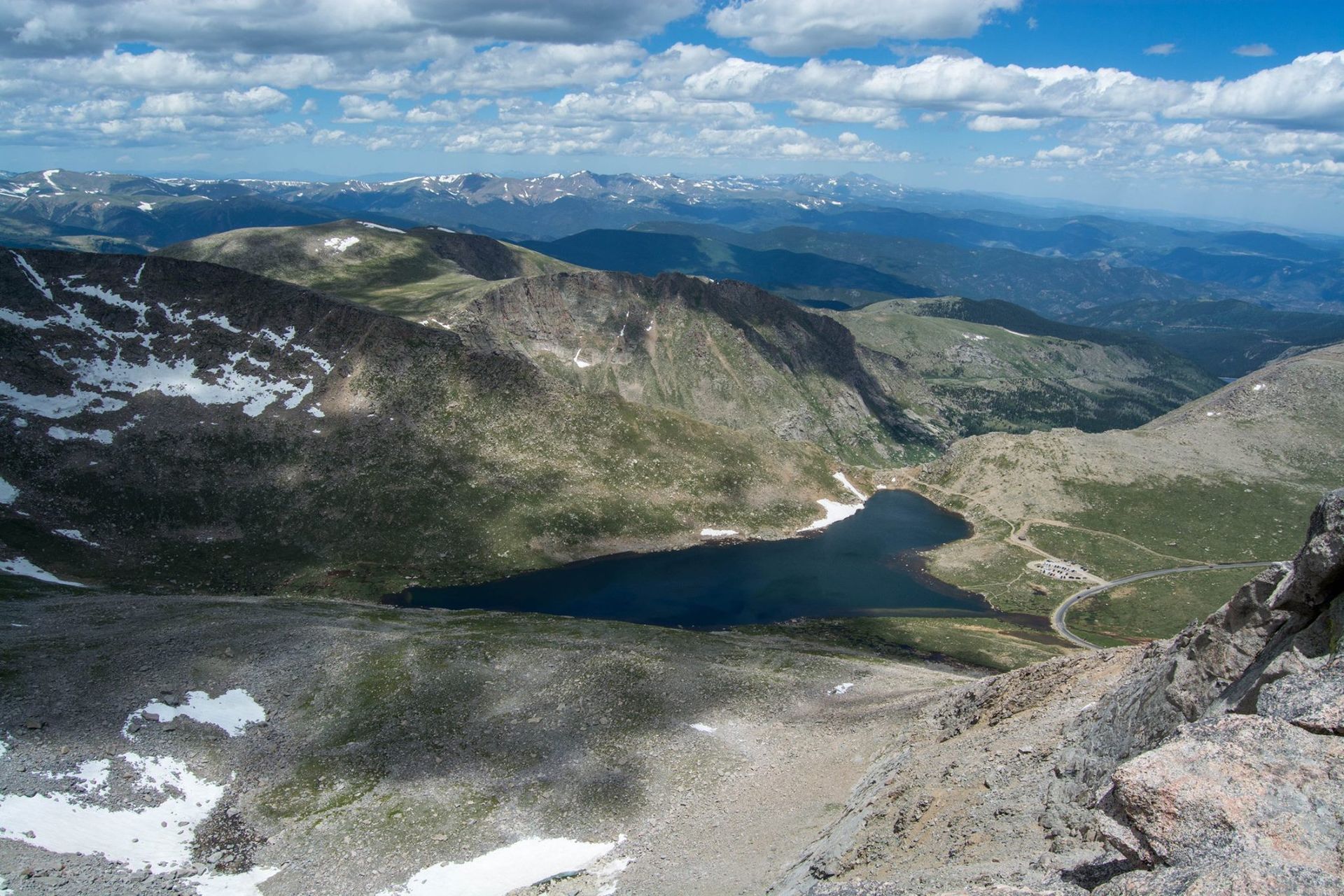 A high-angle view of a mountain valley with a lake. White clouds and snow patches. Green trees and rocky peaks.
