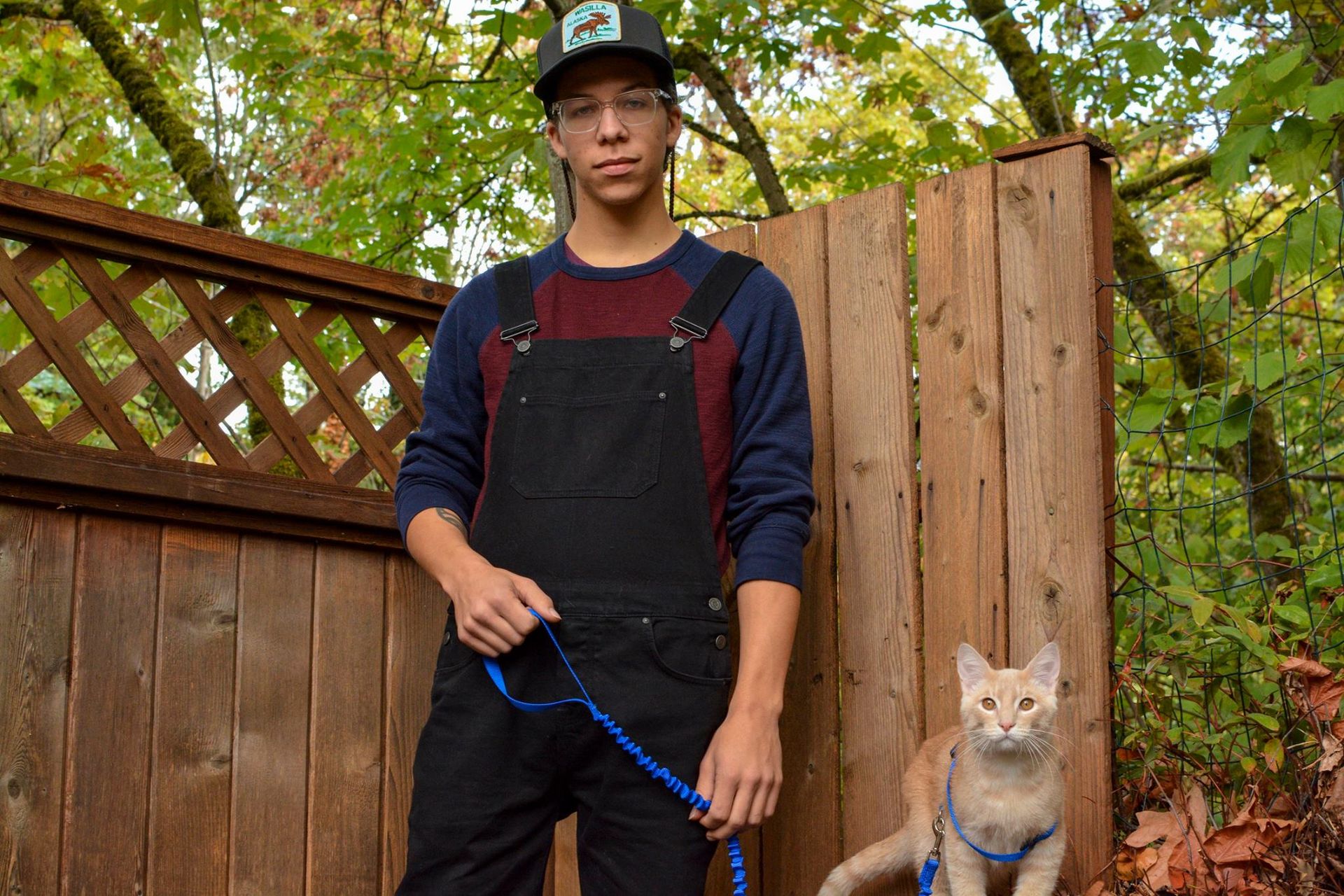Man in overalls and hat, holding cat on leash near wooden fence in fall foliage.