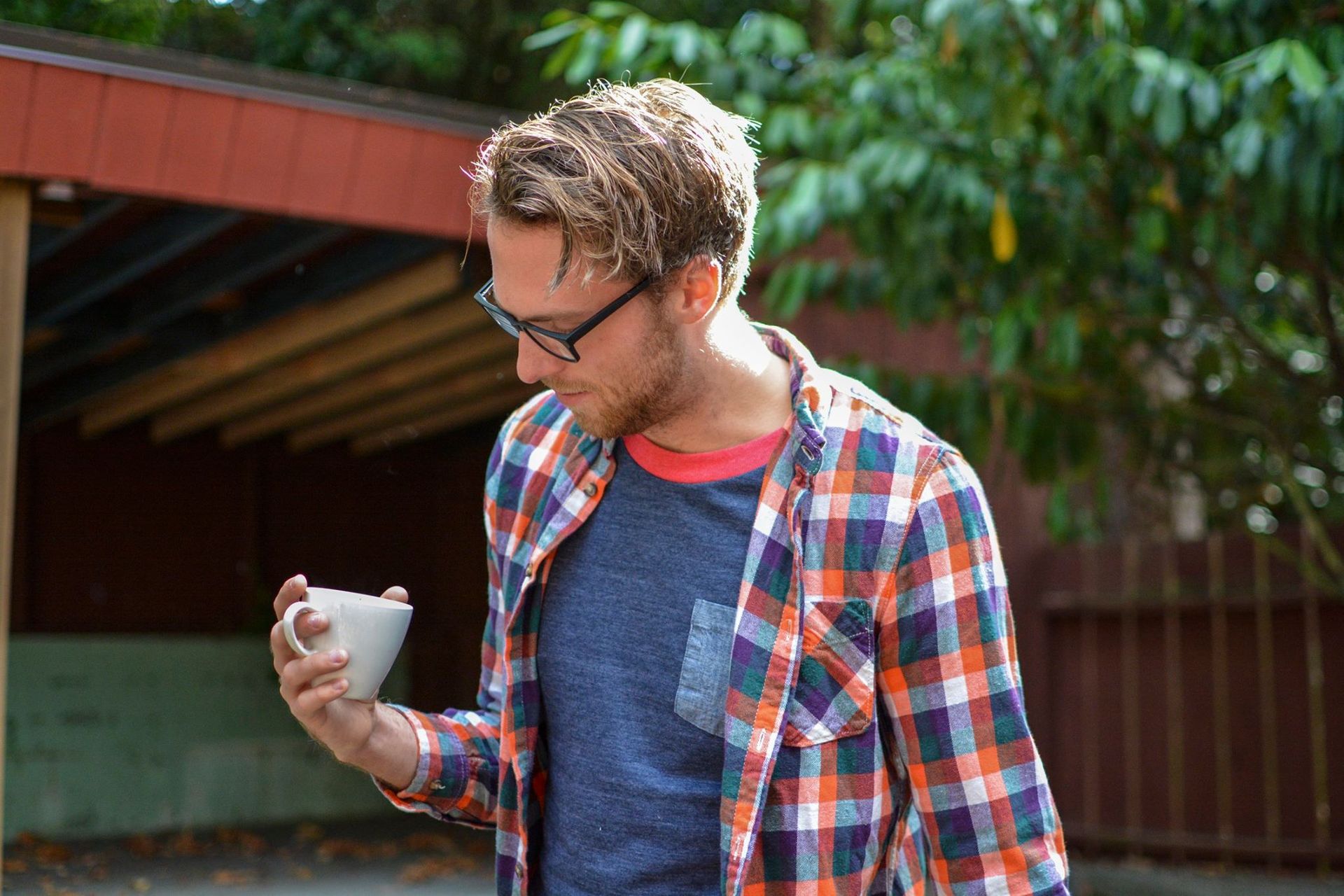 Man in glasses looks at a small white bowl. He wears a plaid shirt, blue shirt, and stands outdoors.