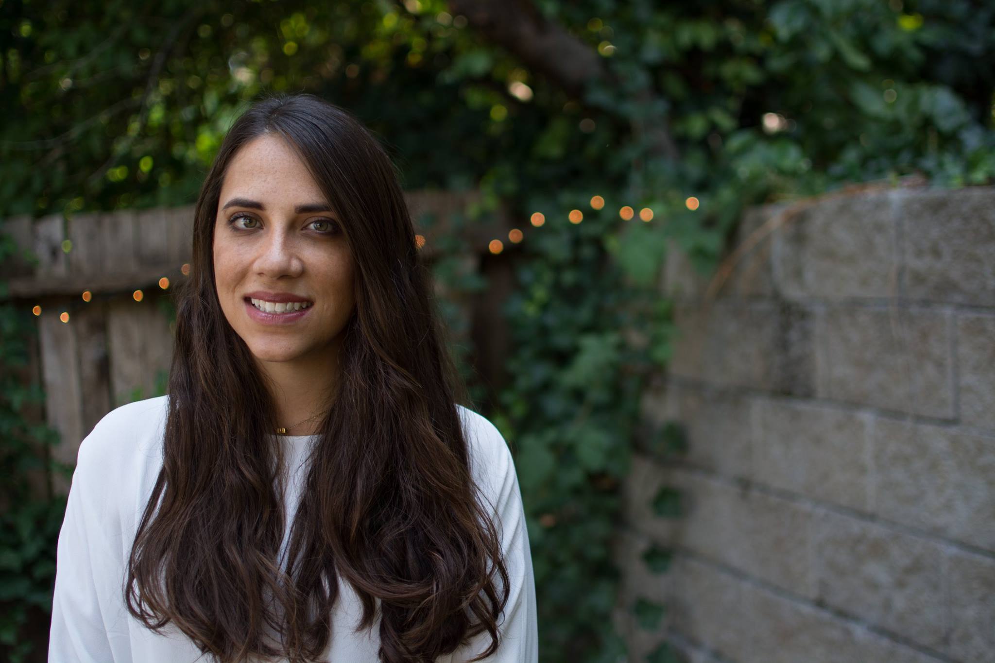 Woman with long dark hair smiles outdoors, in front of a stone wall and foliage.