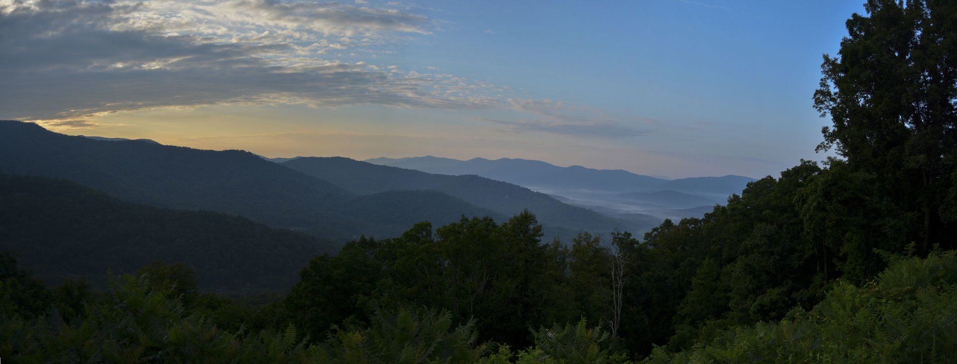 Panoramic view of layered mountain ridges under a blue sky, partially obscured by clouds and mist, with lush green trees in the foreground.