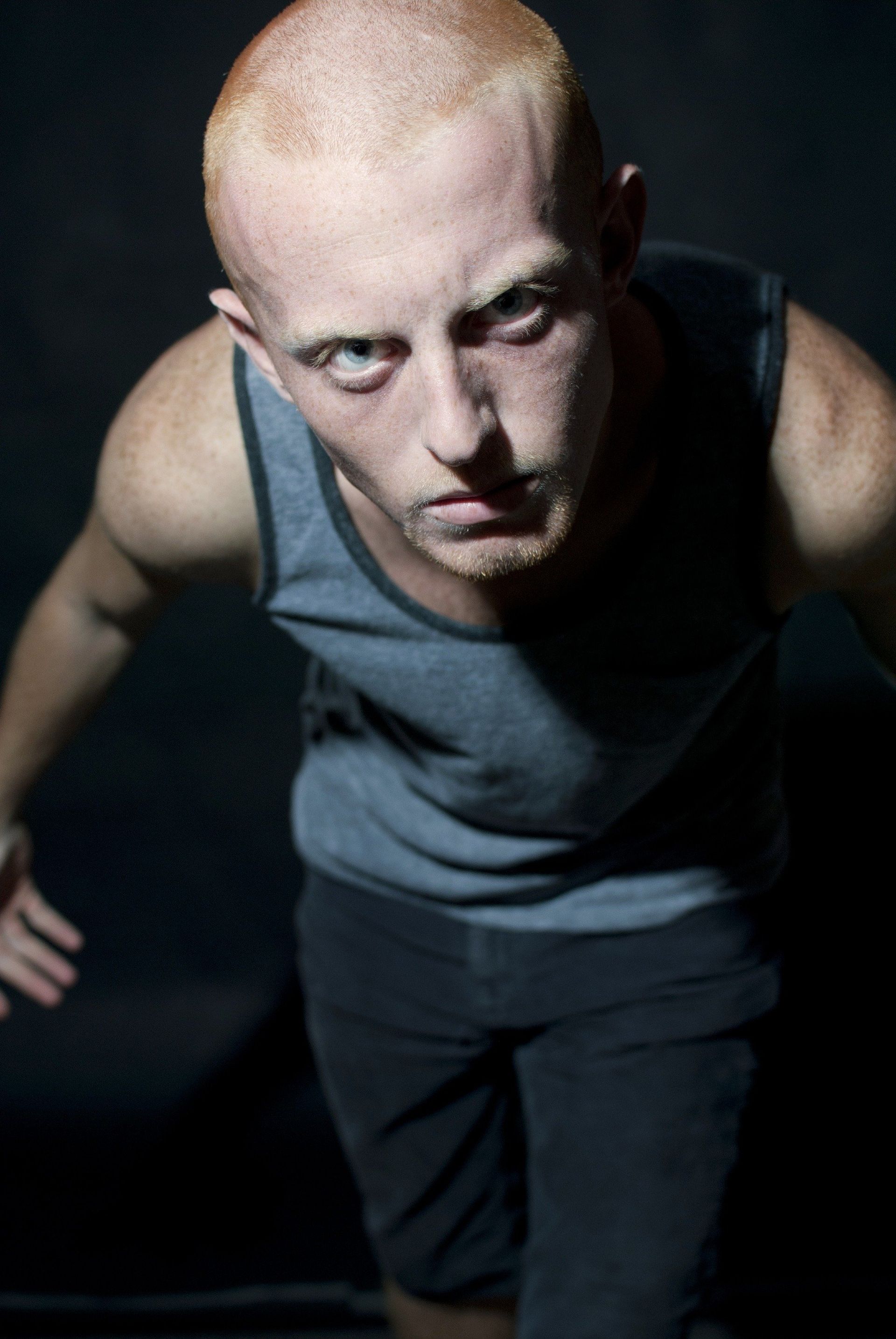 Man with shaved head, gray tank top, and black shorts, looking intensely at the viewer with arms out.
