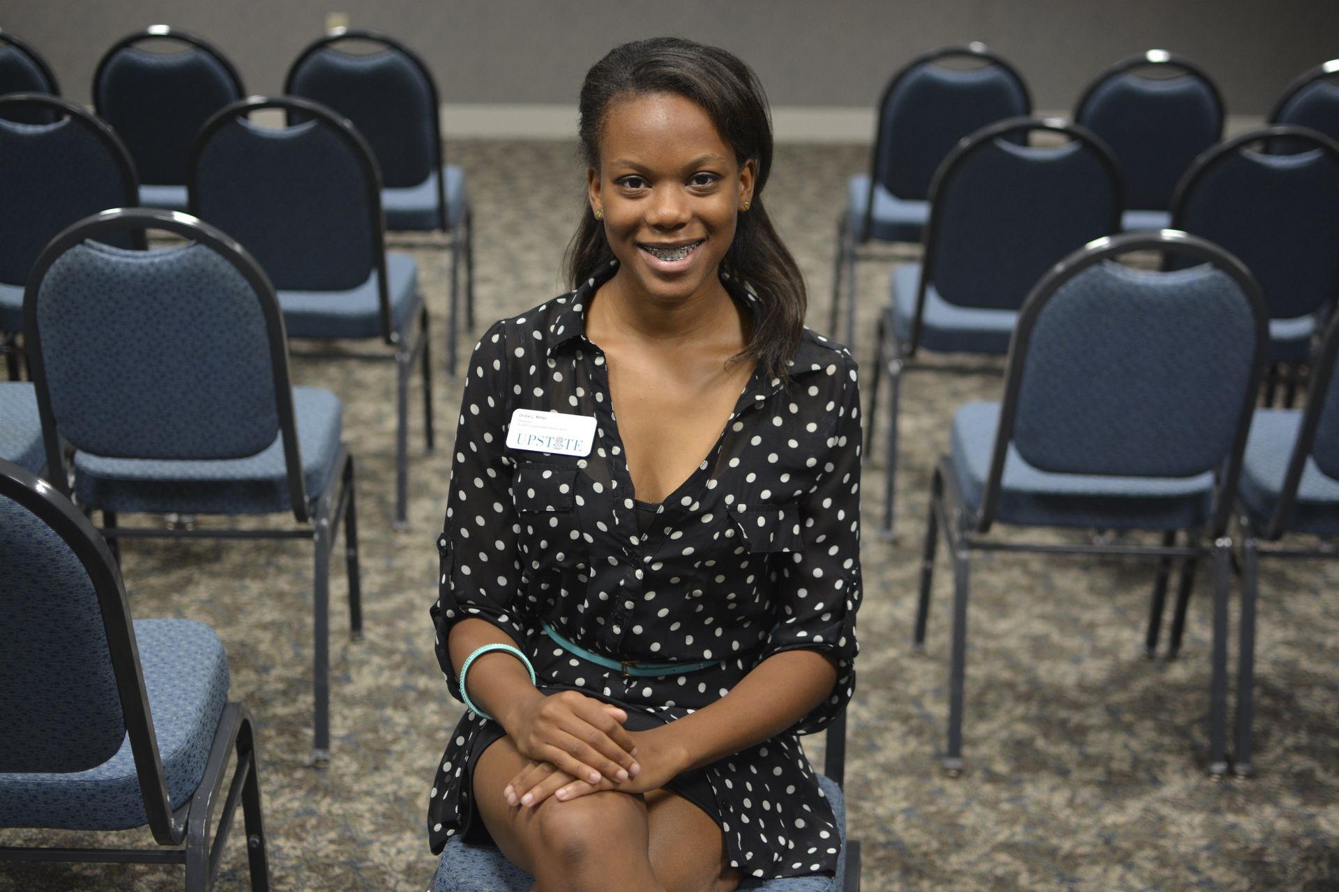 Woman in black patterned shirt smiles while sitting in a room of empty blue chairs.