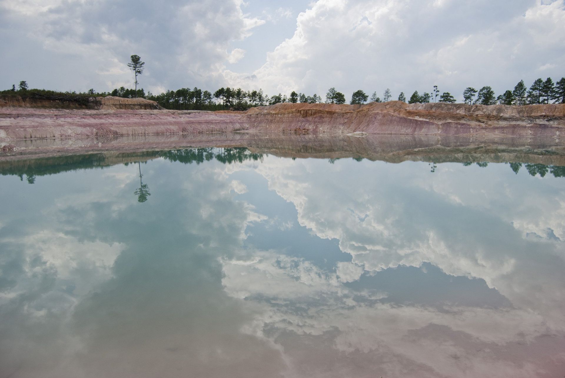 Reflective water surface mirroring a cloudy sky, with a treeline and tower visible in the distance.