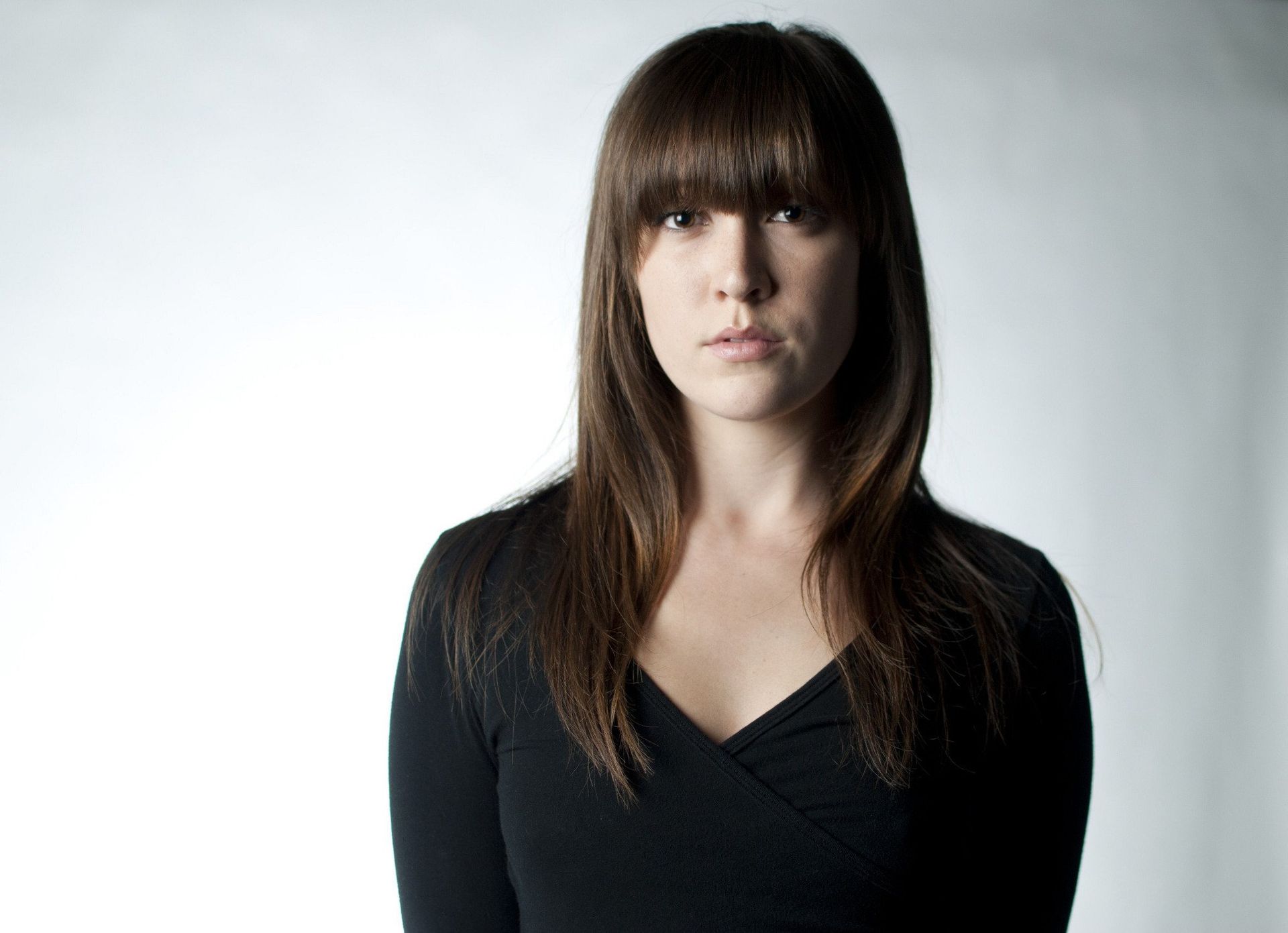 Woman with long brown hair and bangs wearing a black top, staring at the camera.