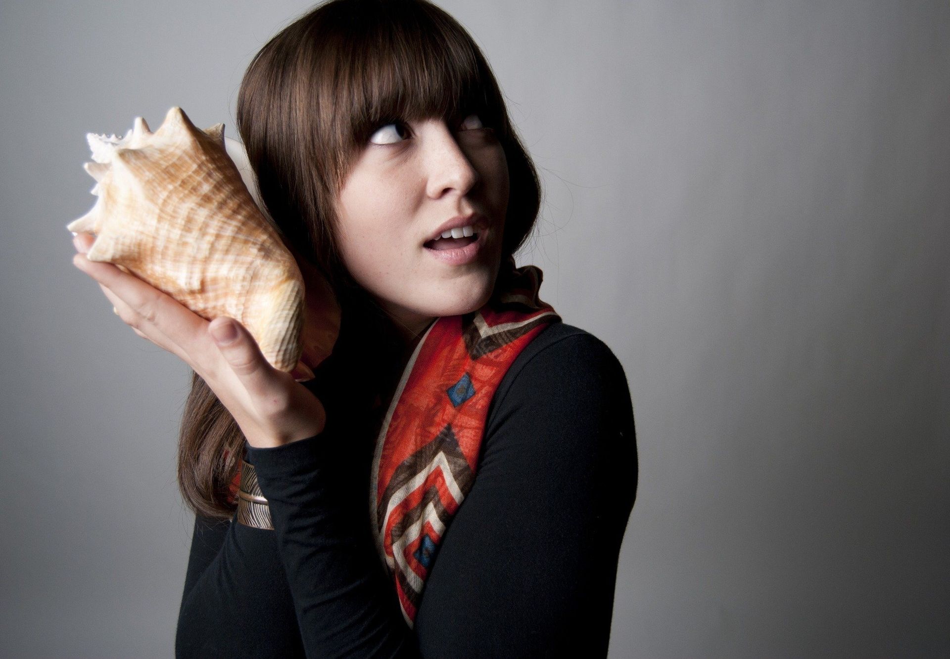 Woman holding a seashell to her ear, looking up with a surprised expression, wearing a black shirt and patterned scarf.
