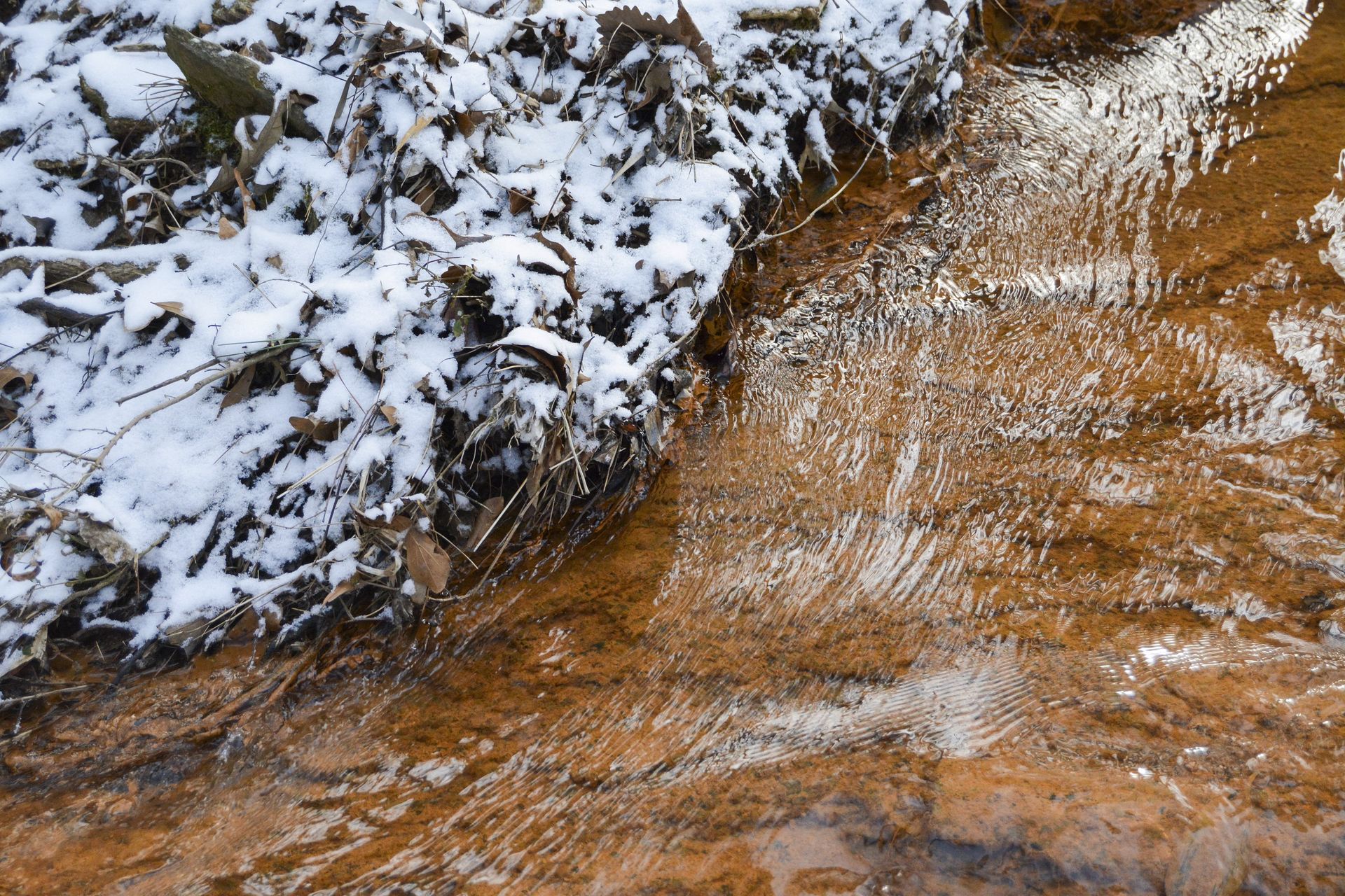Snowy bank borders a shallow stream with brown water.