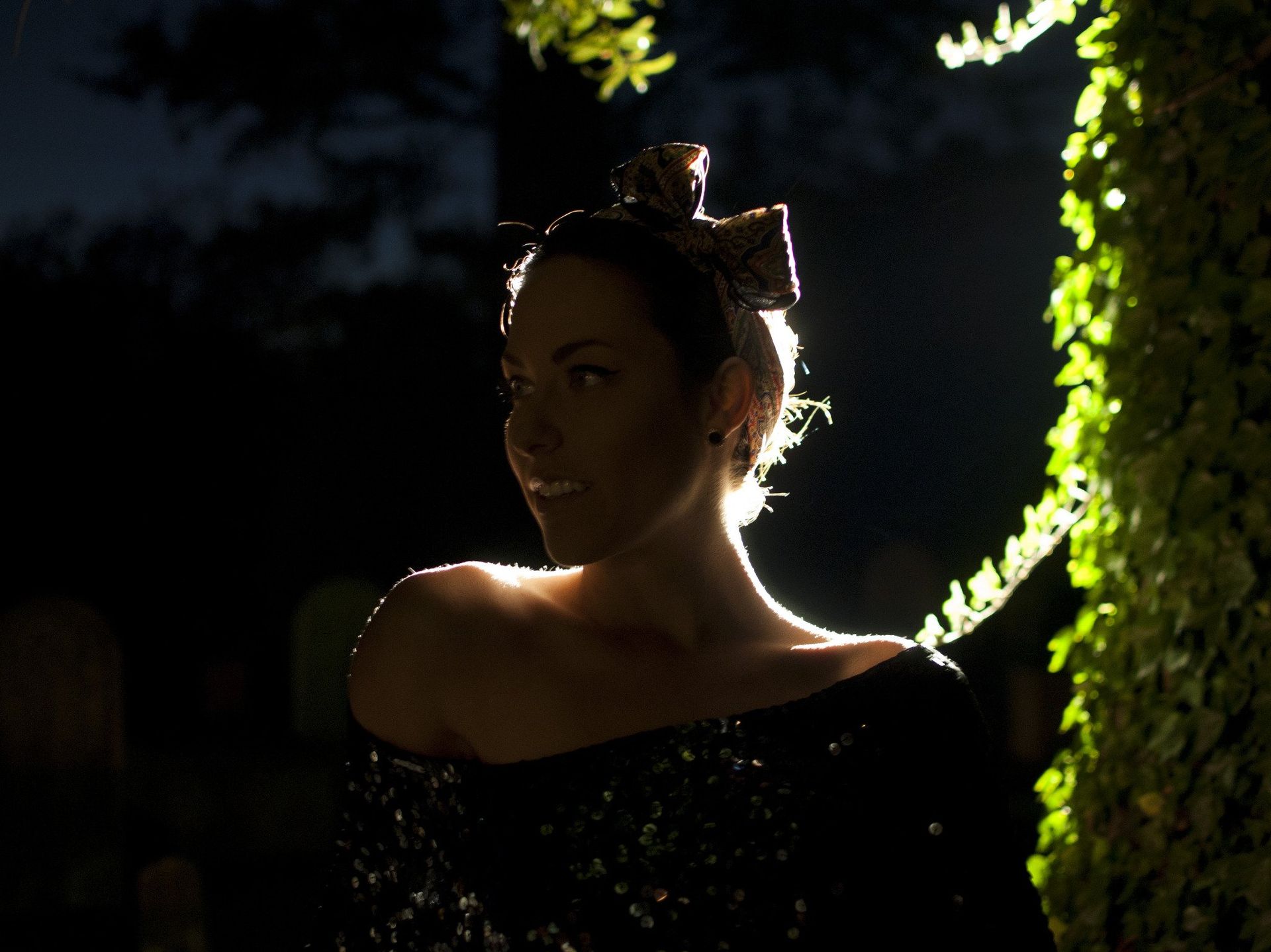 Woman smiling, illuminated by backlight. Wearing a dark dress and headpiece, next to greenery.