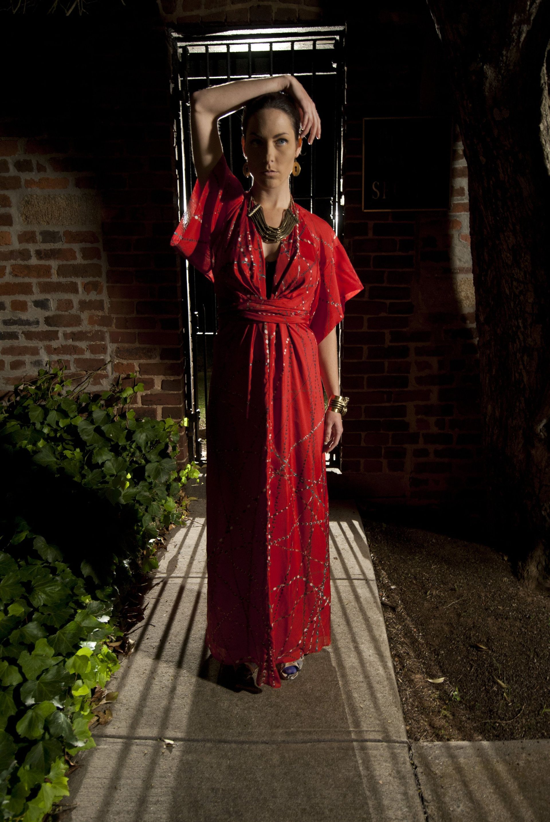Woman in red sequined dress poses outdoors, hand on head, near a gate.