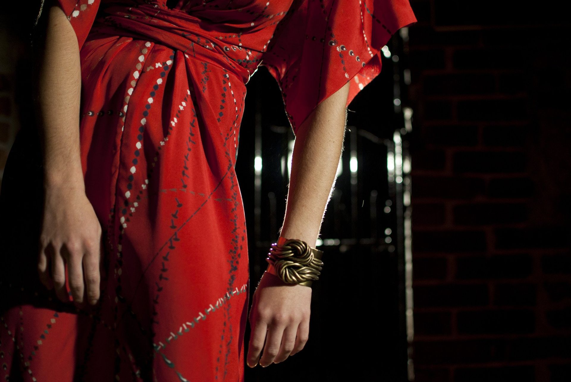 Woman in a flowing red dress, with gold bracelet, stands in a dark doorway.