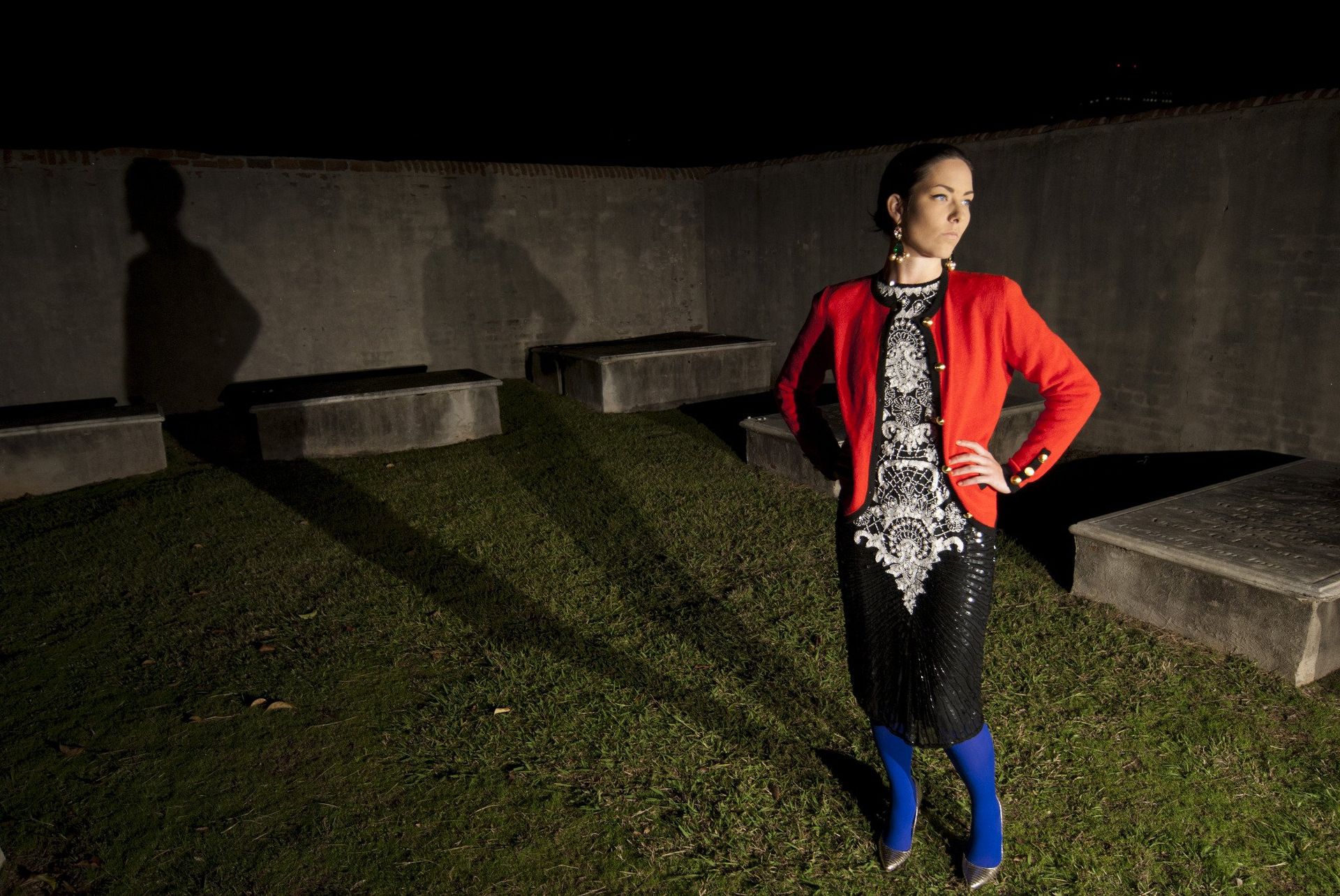 Woman in red jacket and patterned top stands on grass with shadow in front of concrete wall.