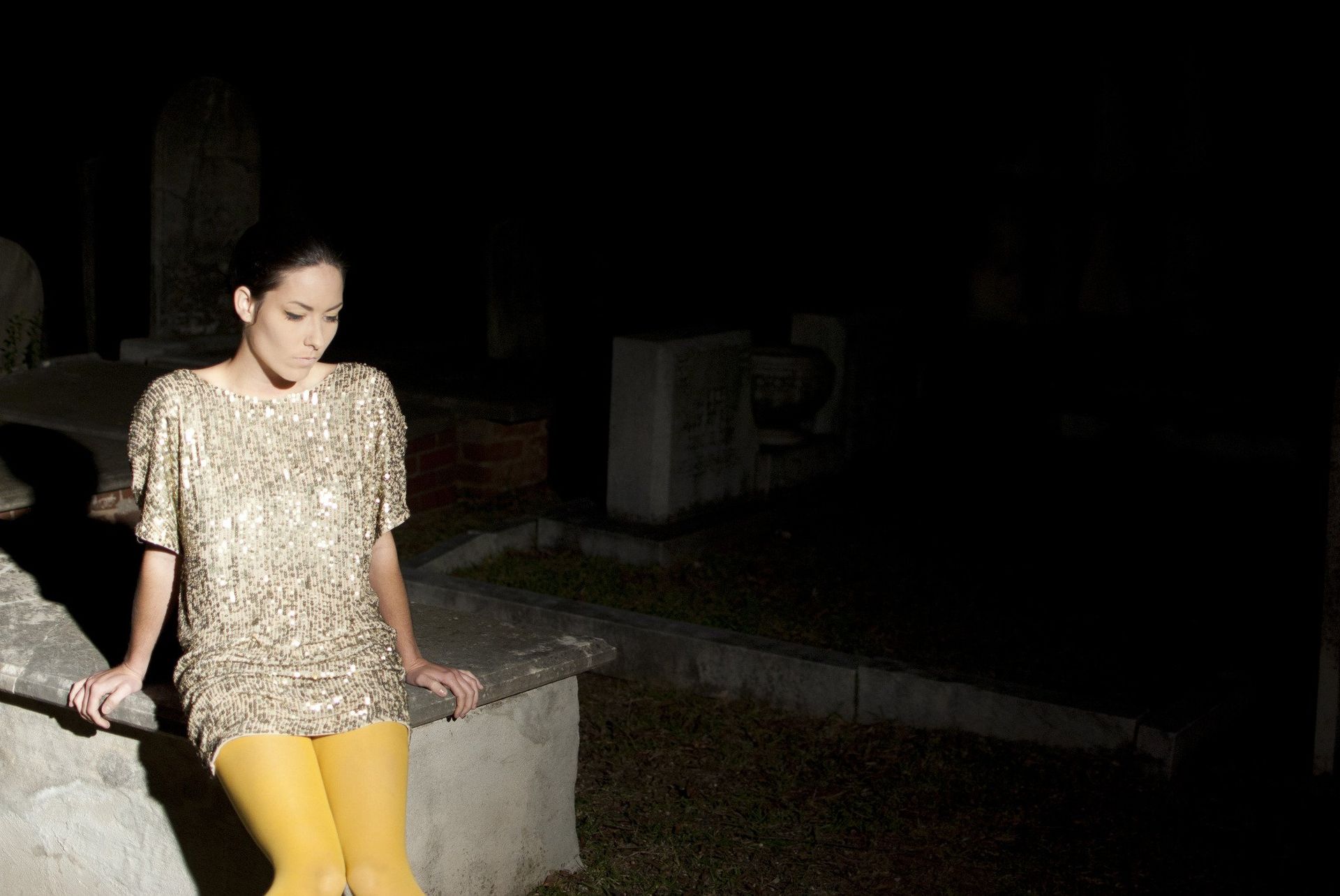 Woman in gold dress and yellow leggings sits on tombstone at night.