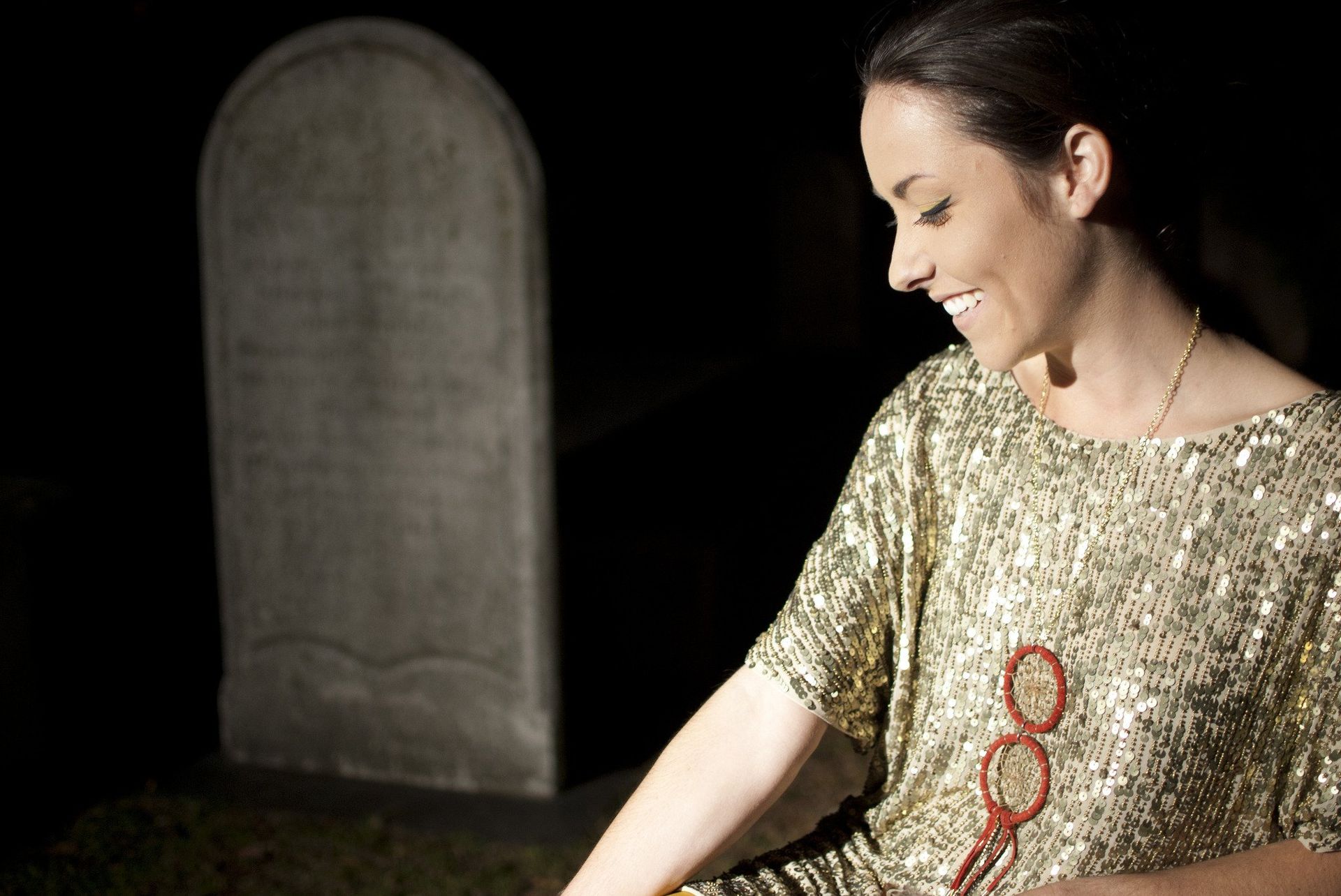 Woman in sparkly gold top smiles near a tombstone at night.