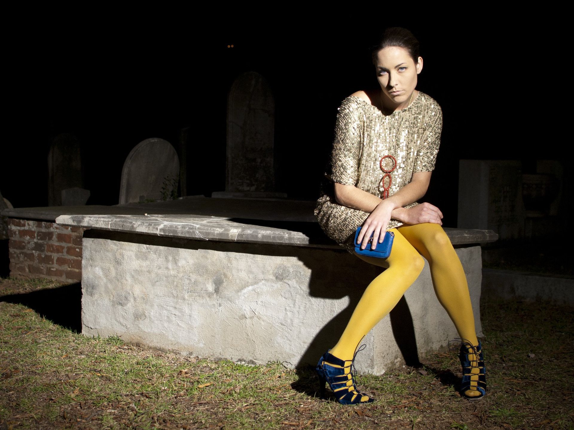 Woman in gold dress, yellow tights, and heels sits on tomb in a dark cemetery.