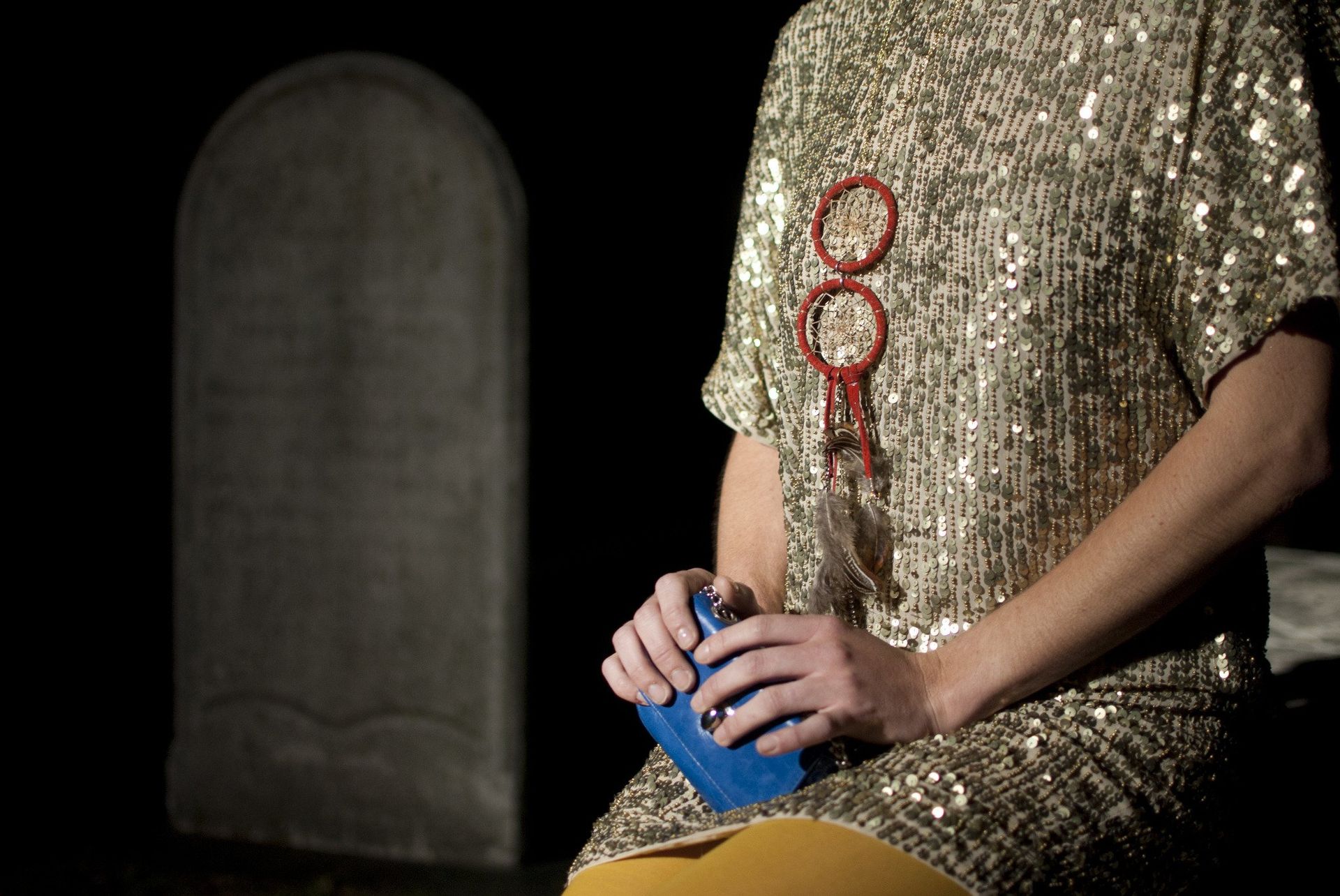 Person in gold sequined dress holds blue object near tombstone. Dark setting.