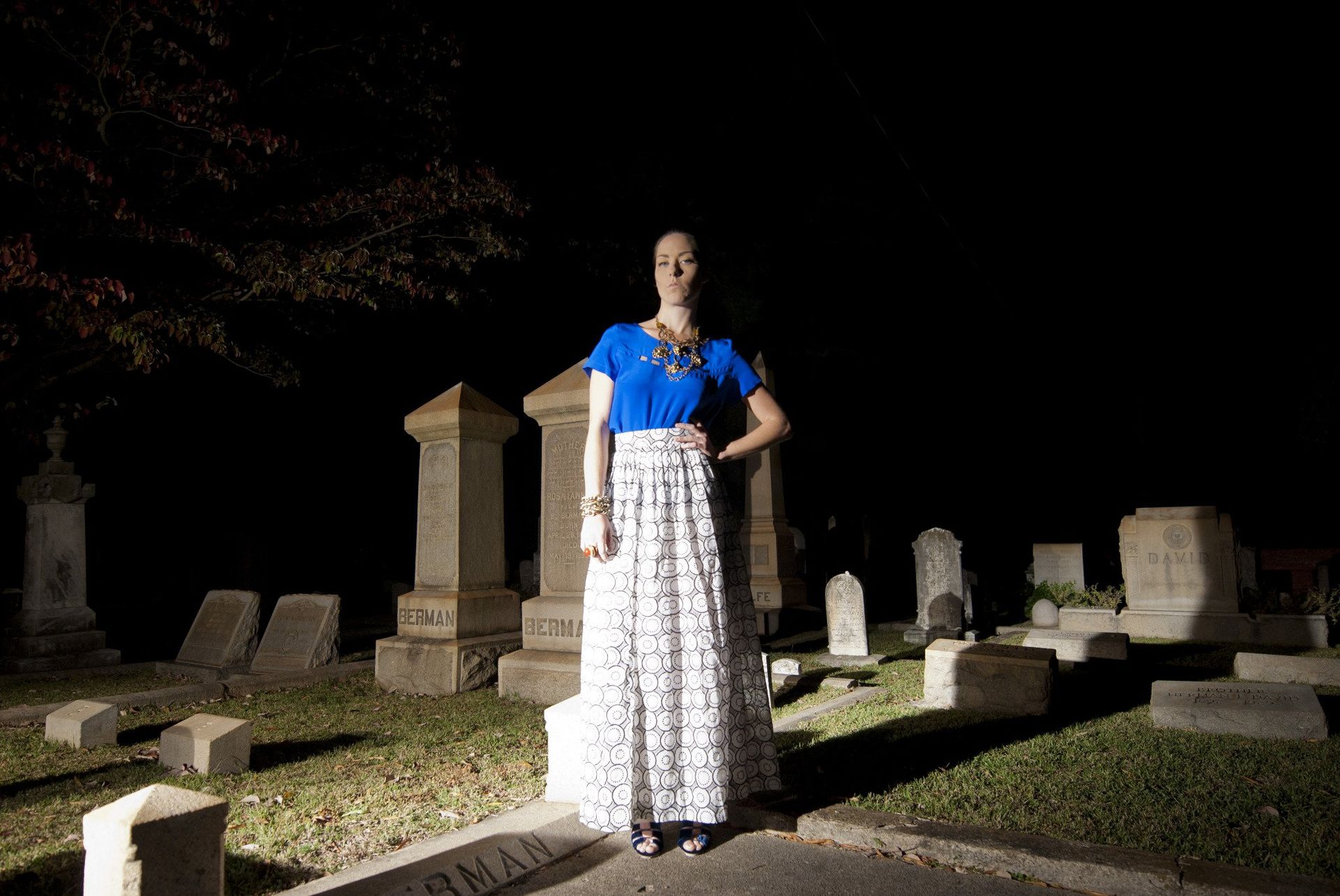 Woman in blue top and long skirt stands in a graveyard at night, lit by a spotlight.
