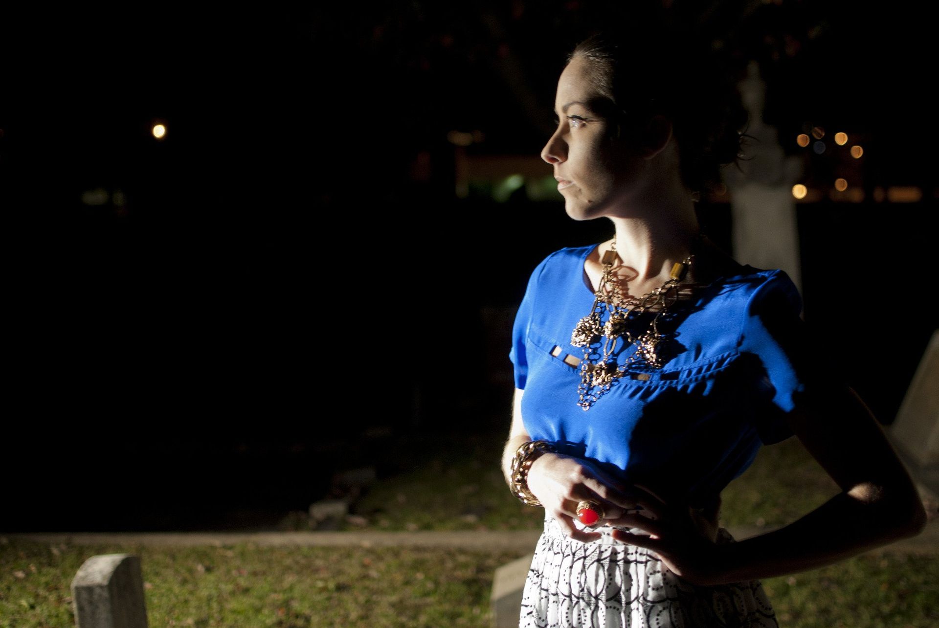Woman in blue top, white patterned skirt, looking to the side in a dark cemetery.