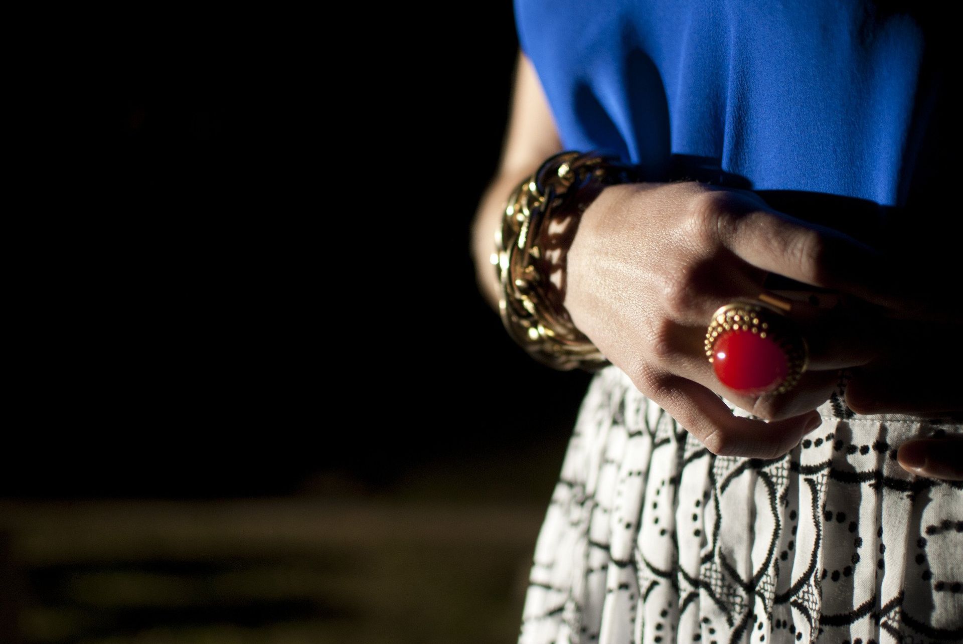 Woman's hand with gold bracelet and ring. Blue top, patterned skirt. Dark background.