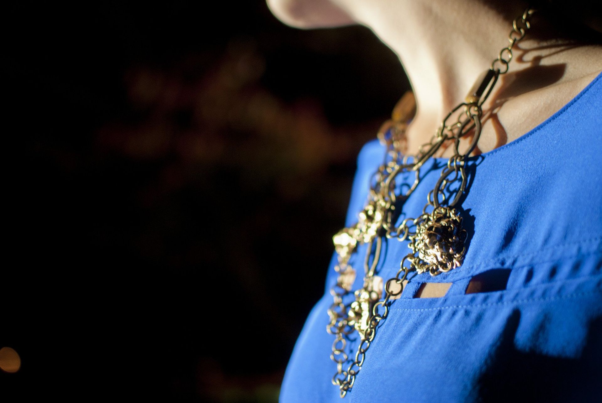 Woman wearing a gold necklace over a blue top, against a dark background.