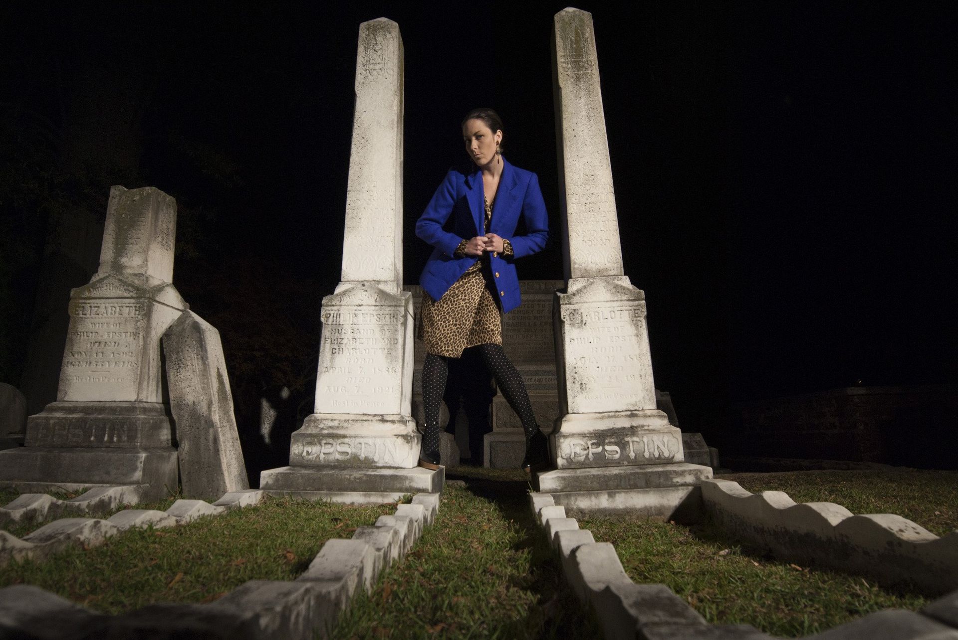 Woman in blue blazer stands between two tall, white stone pillars in a dark cemetery.