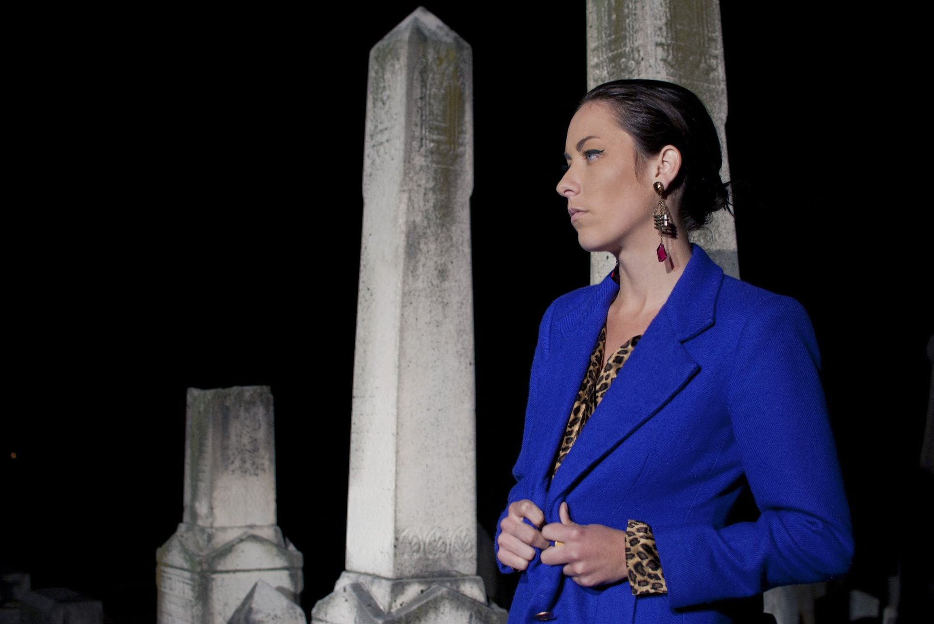 Woman in blue jacket at a cemetery at night, looking to the side, near stone pillars.