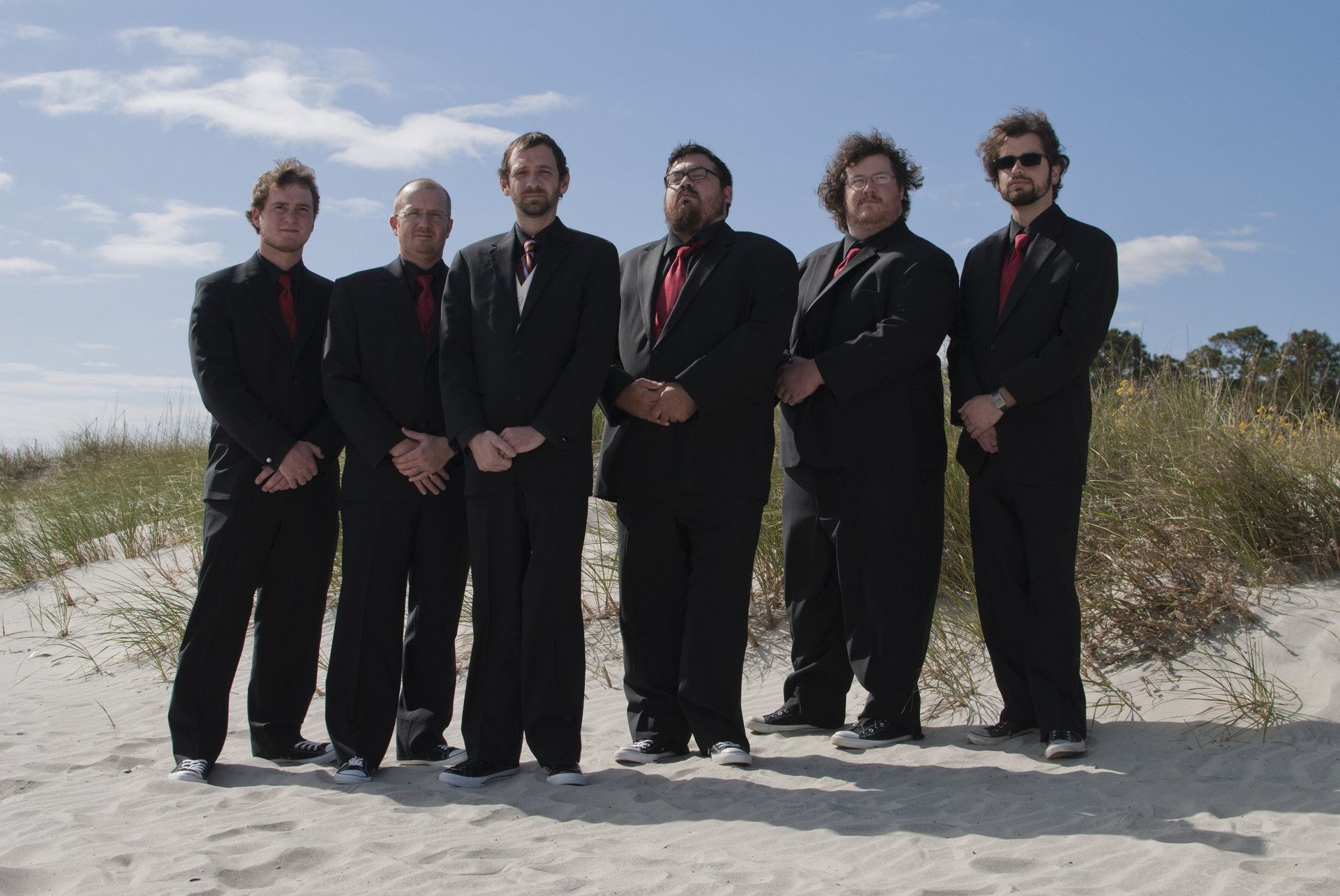 Six men in black suits stand on a beach, blue sky overhead.