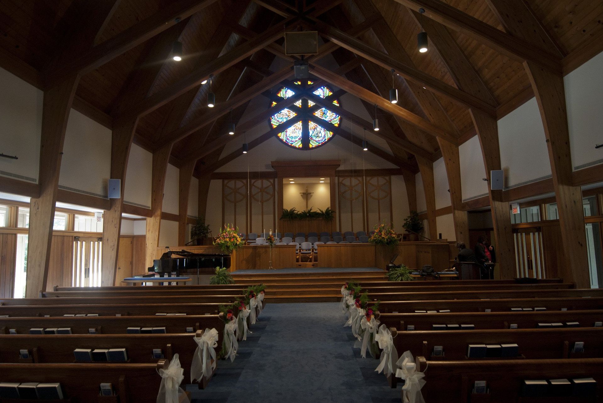 Interior of a church with rows of pews, a stage, and a stained-glass window.