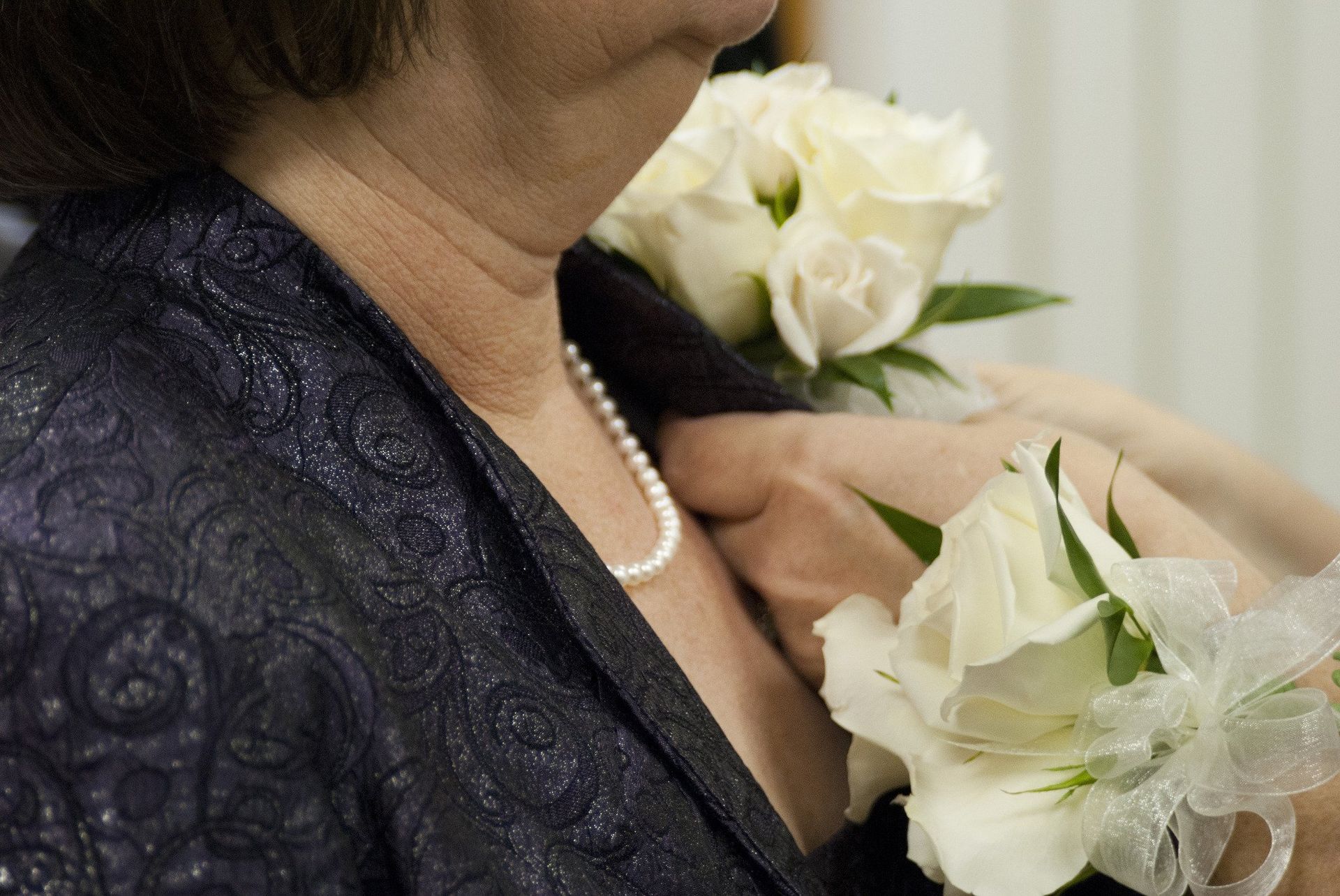 Woman in dark patterned jacket with a white flower corsage adjusting her pearls.
