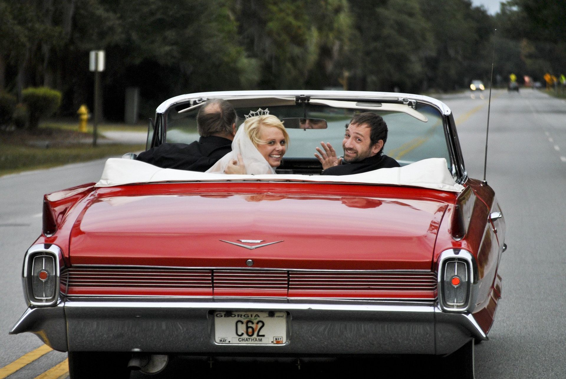 Bride and groom wave from a red convertible; they drive down a road after a wedding ceremony.