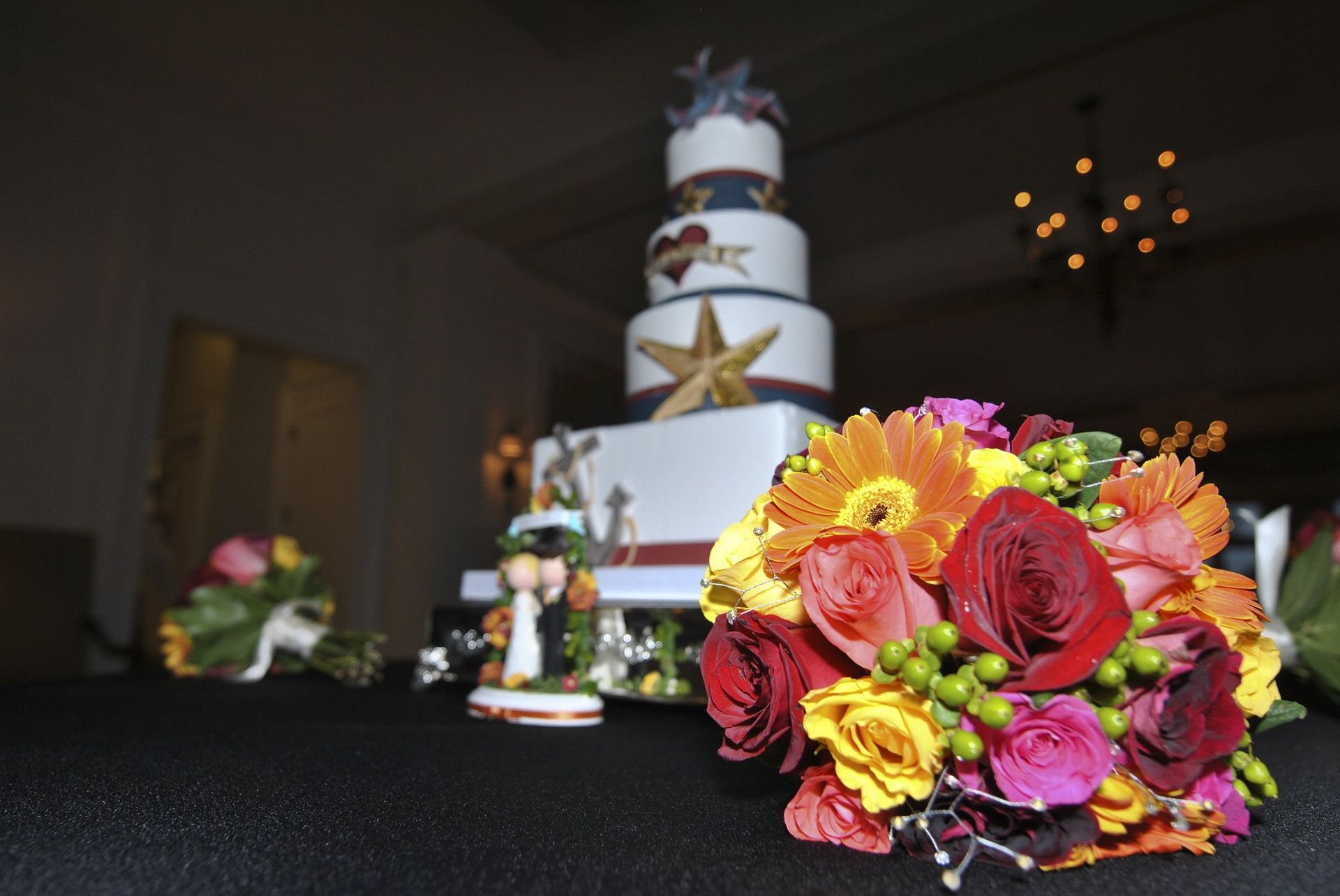 Wedding cake and colorful bouquet on a black table; blurred figures and chandelier in the background.