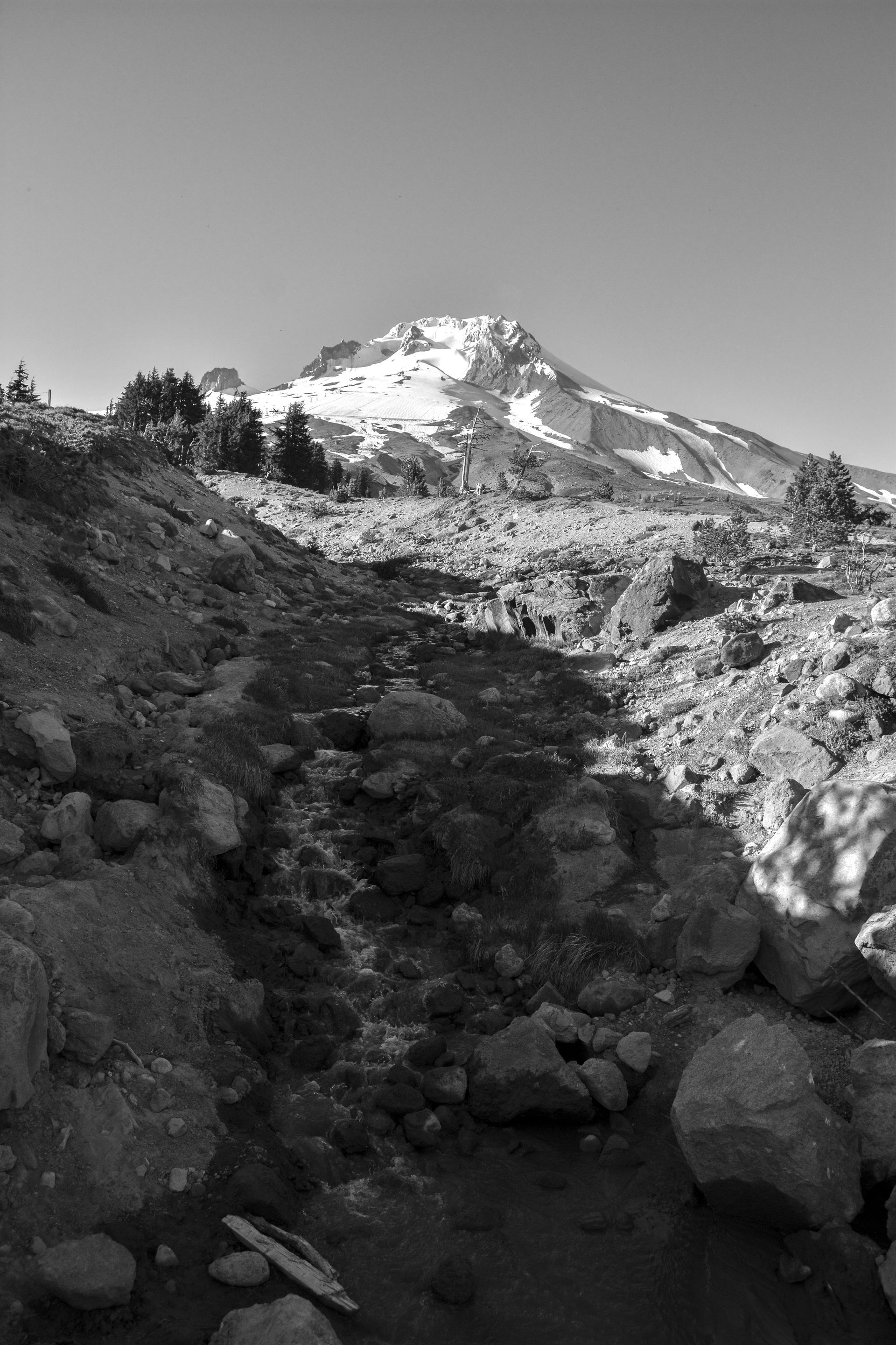 Black and white view of a mountain with snow, rocky terrain, and a stream flowing in the foreground.