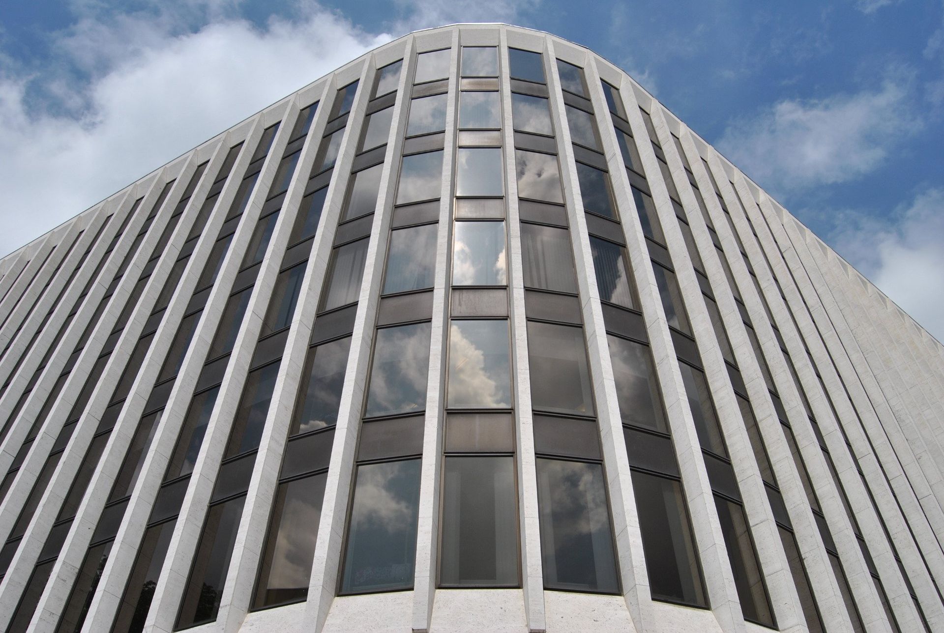 White office building with vertical ribs and corner windows, against a cloudy sky.