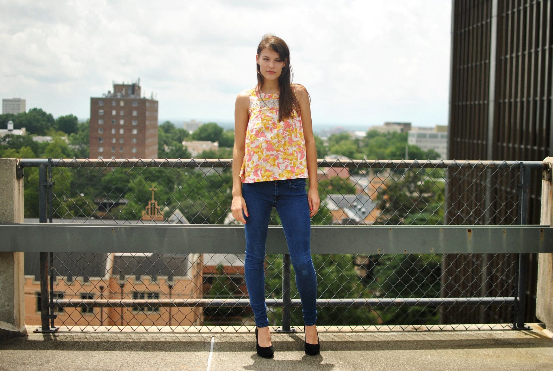 Woman in floral top and jeans stands on a rooftop overlooking a city.