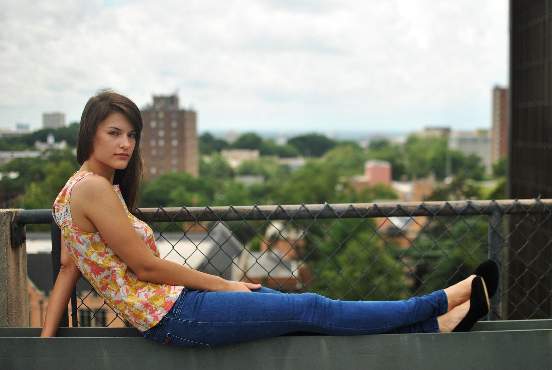 Woman lounging on a rooftop ledge; city view in background, wearing jeans and floral top.