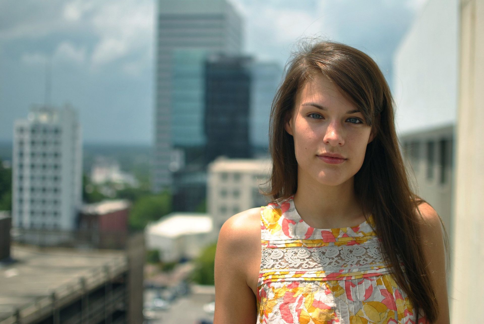 Woman with long brown hair, wearing a floral dress, smiles slightly outdoors with city buildings in background.