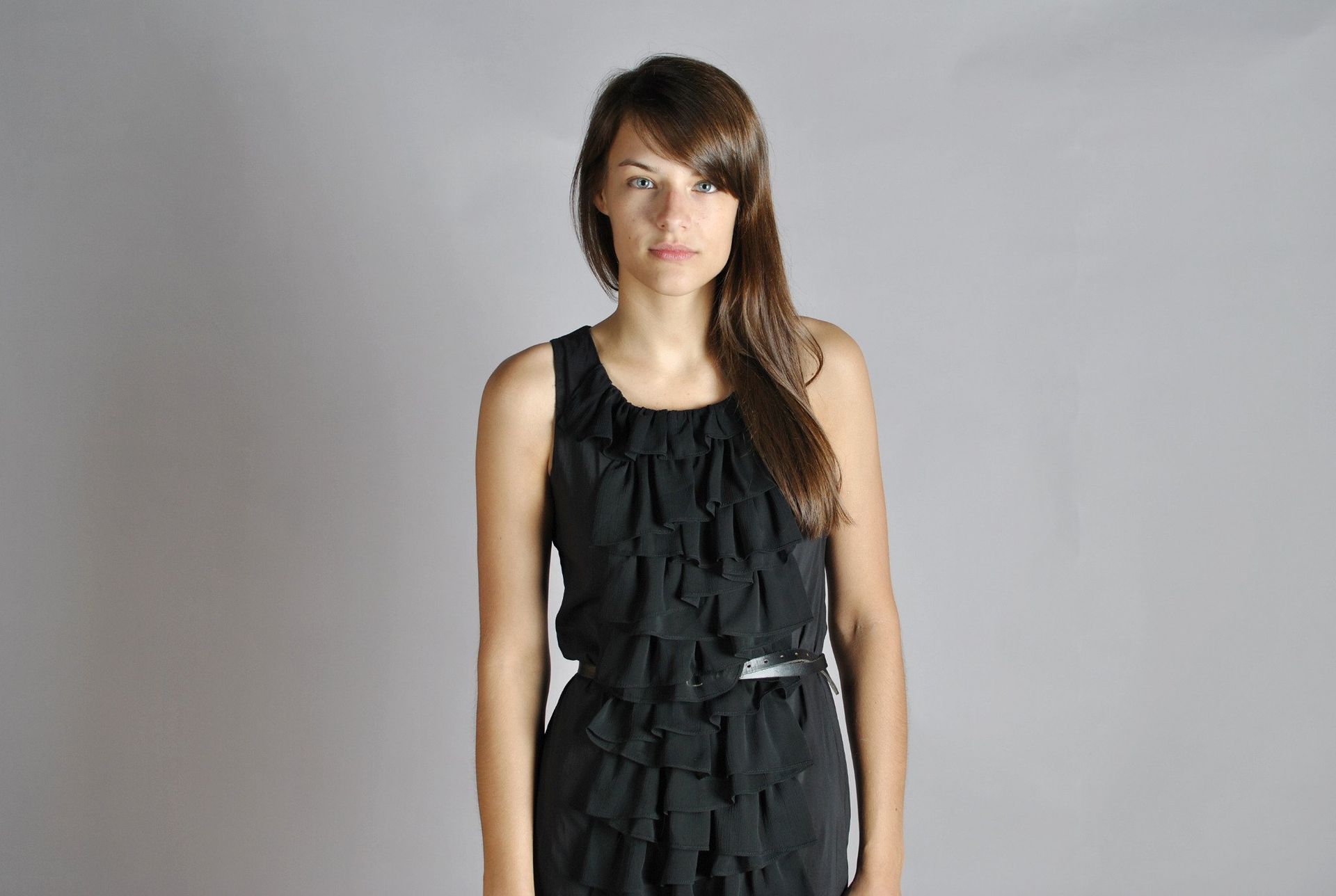 Woman in a black dress stands facing forward against a gray backdrop. The dress has ruffles and a belt.