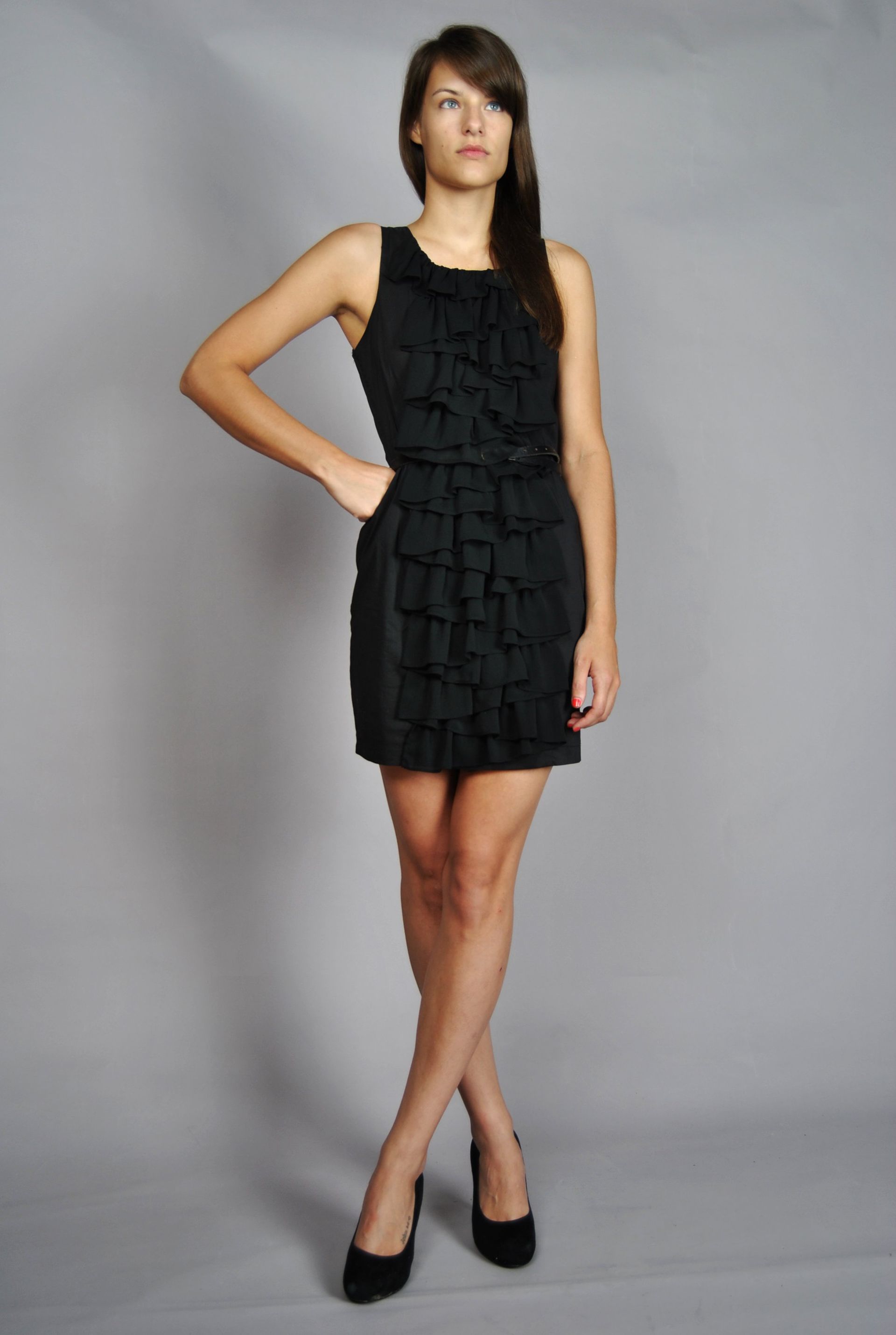 Woman in black dress, ruffles detail, posing with hand on hip. Crossed legs, heels.