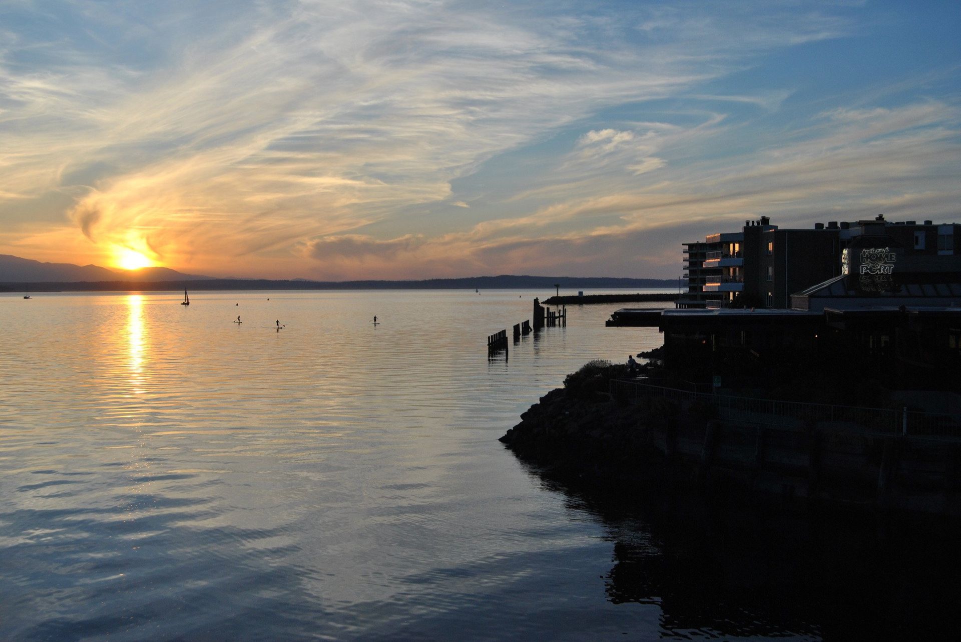 Sunset over calm water; silhouetted shoreline and buildings. Sky features wispy clouds and orange sun.