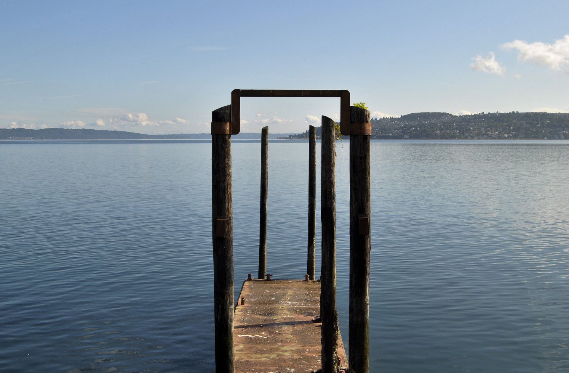 Wooden pier posts extending into calm blue water, hills in the background under a blue sky.