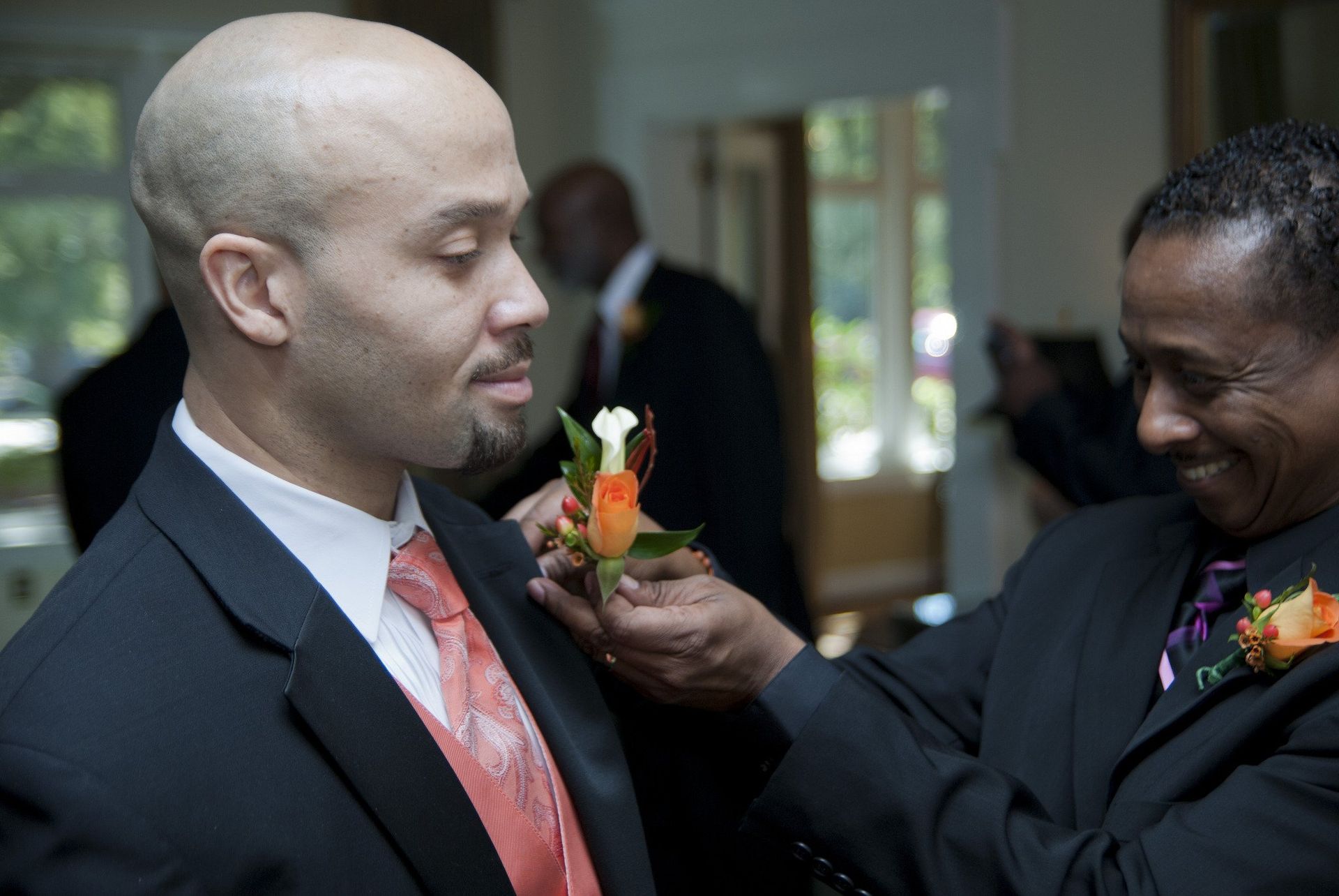 Man in suit has boutonniere adjusted by another man. Indoors, other people in suits are visible.