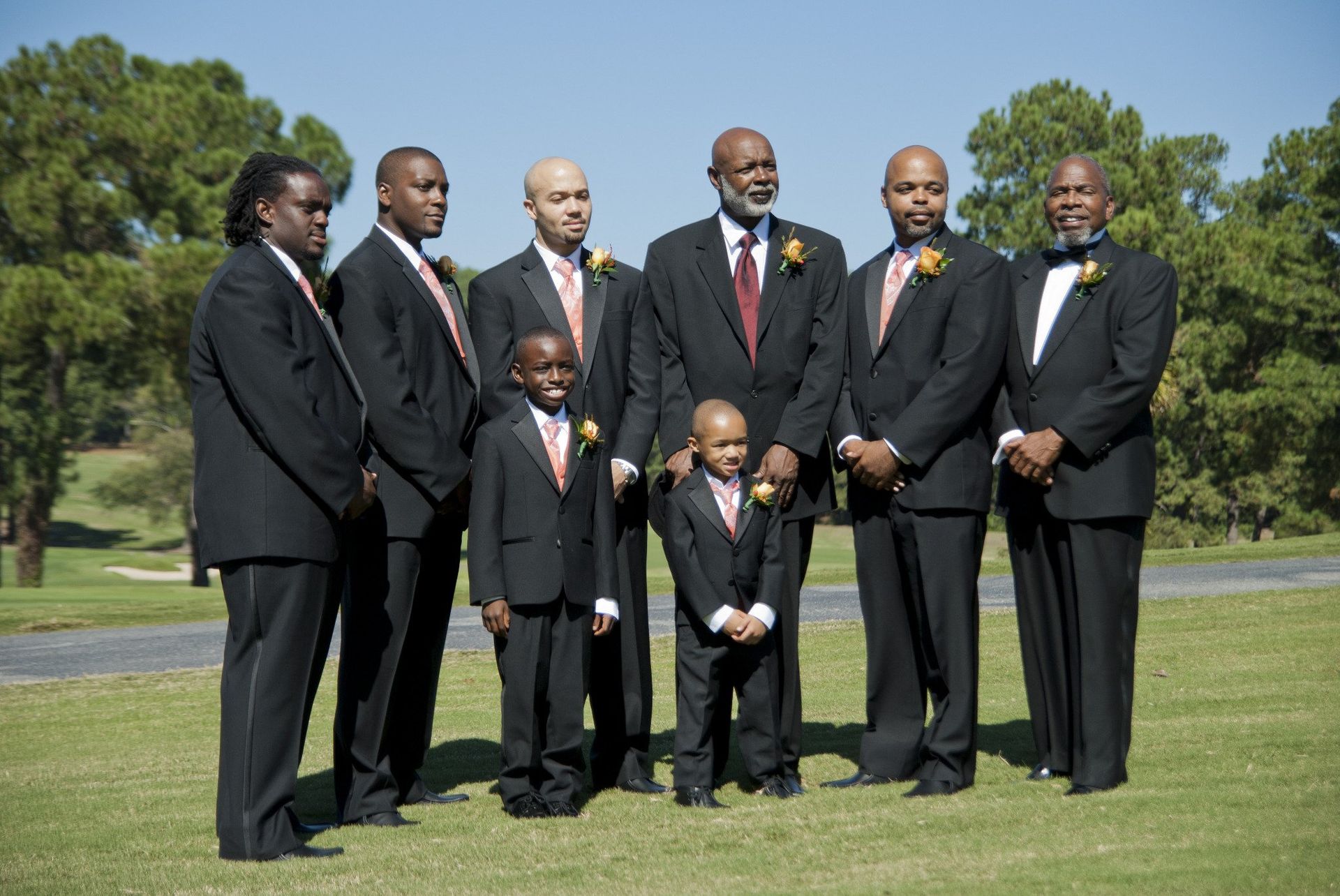Groomsmen in black tuxedos with orange accents, standing on grass. Two children are included.