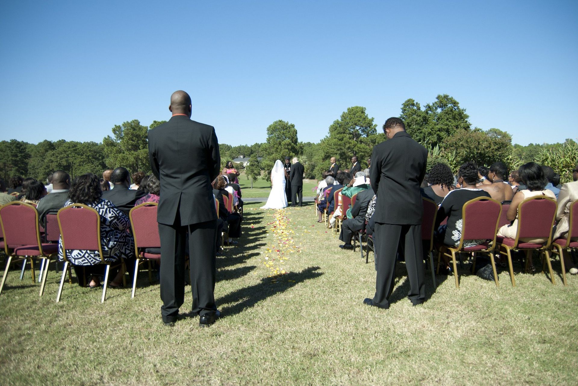 Wedding ceremony outdoors with guests seated, two people in suits, bride and groom at altar.