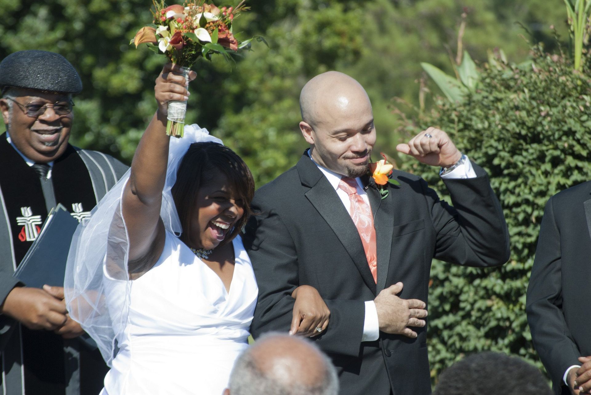 Newly married couple exiting ceremony, bride with bouquet raised, groom smiling; officiant in background.