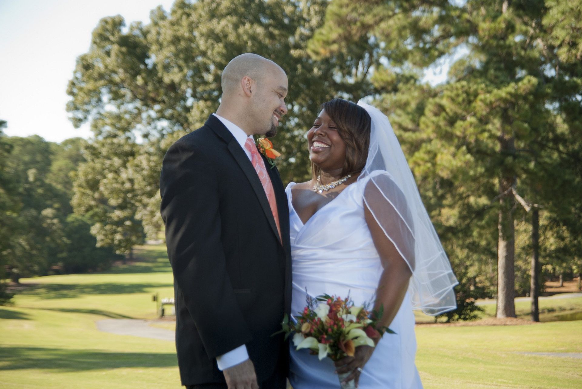 Bride and groom smiling at each other outdoors. Bride in white dress, groom in black suit, holding bouquet. Green background.