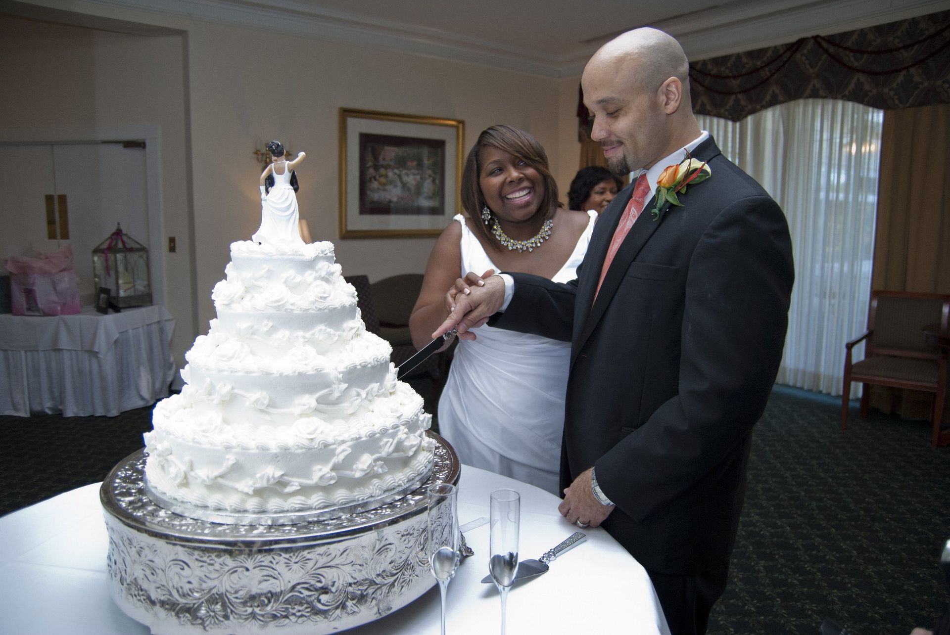 Couple cutting wedding cake at reception. Smiling woman, suited man with orange flower. Formal setting.