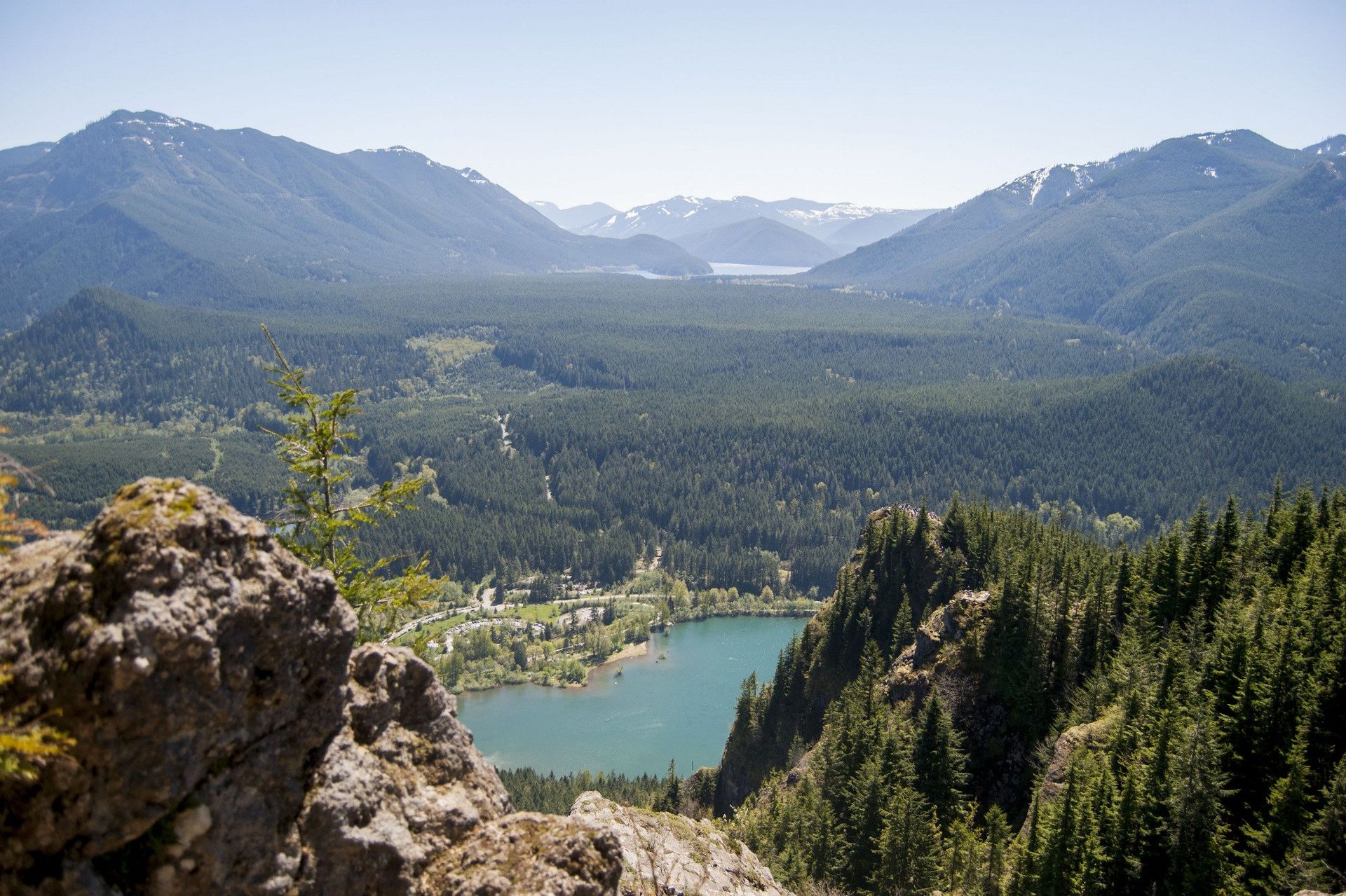 Mountain landscape with a blue lake surrounded by dense evergreen forest and snow-capped peaks.