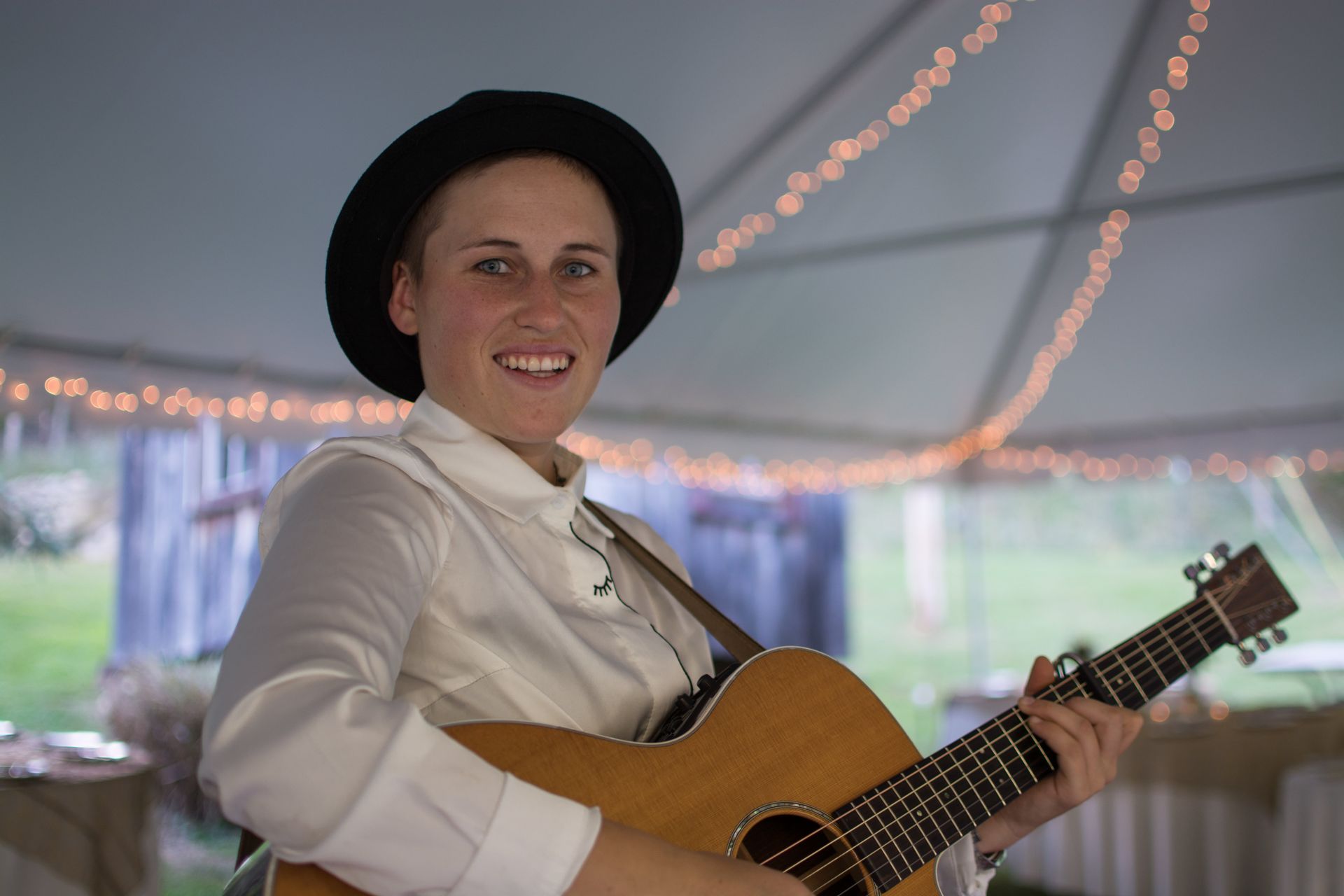 Person wearing a black hat, playing an acoustic guitar, smiles in a tent with string lights.