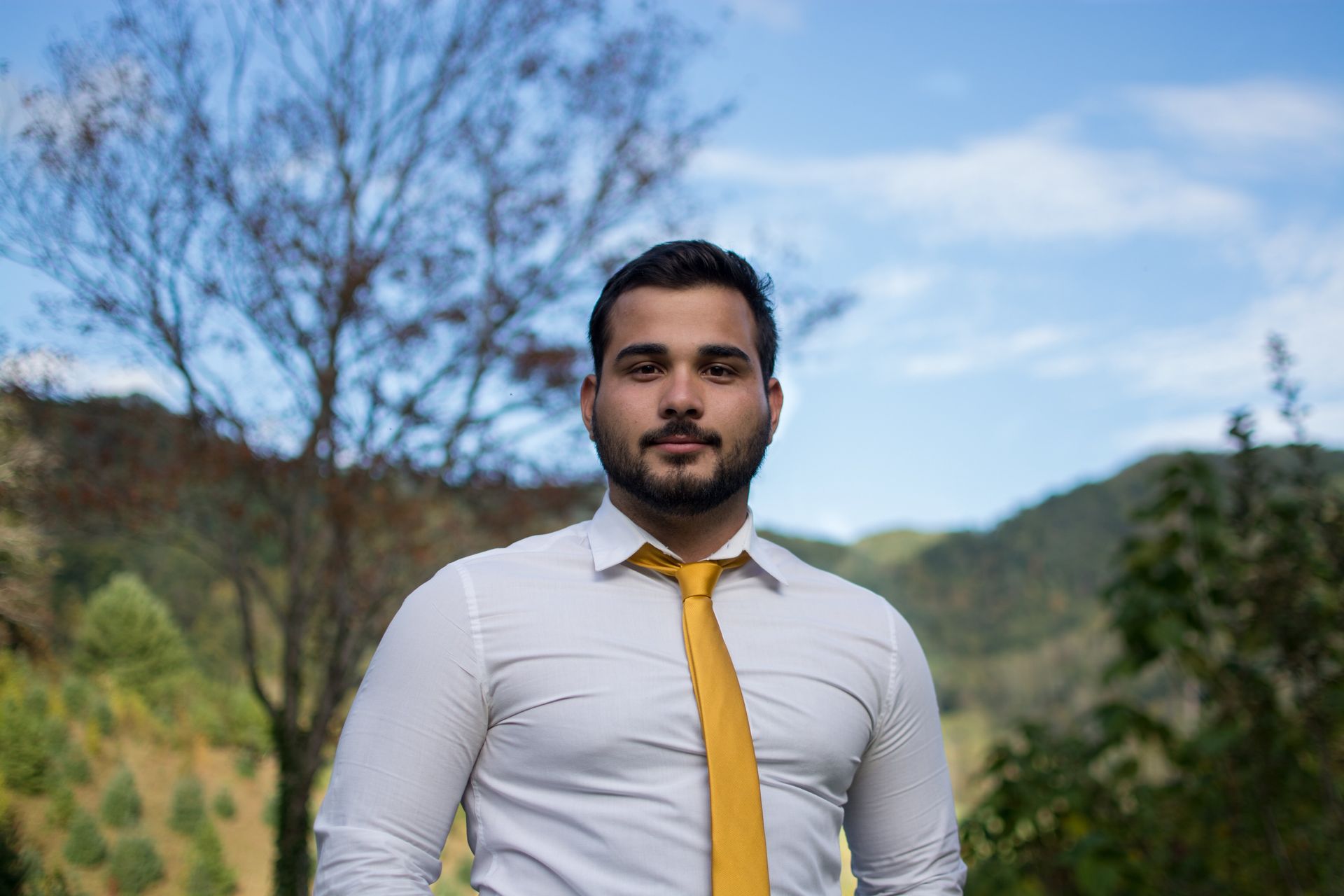 Man in white shirt and yellow tie against a blurry outdoor background of trees and sky.