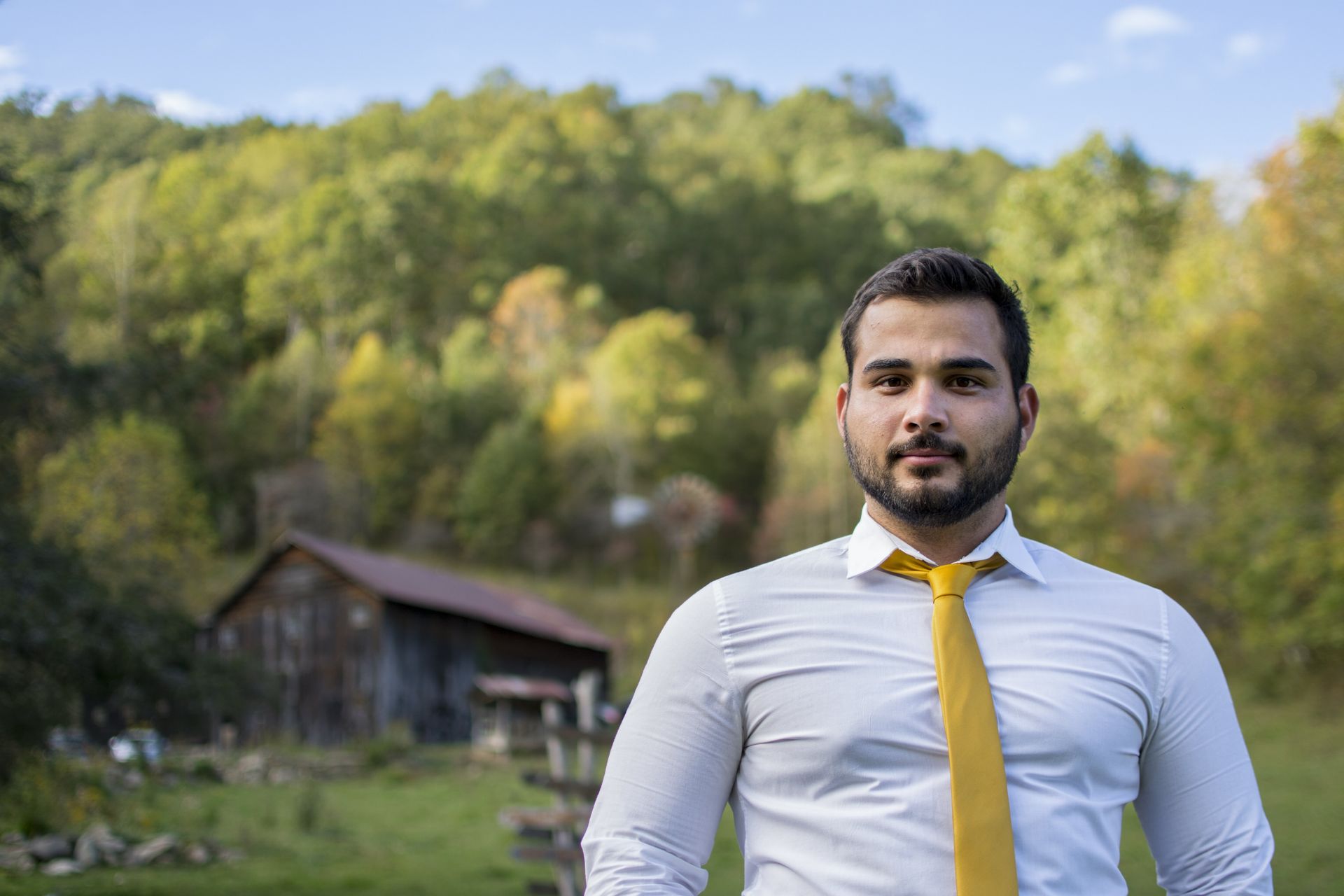 Man in white shirt and yellow tie stands in field with barn and autumn trees.