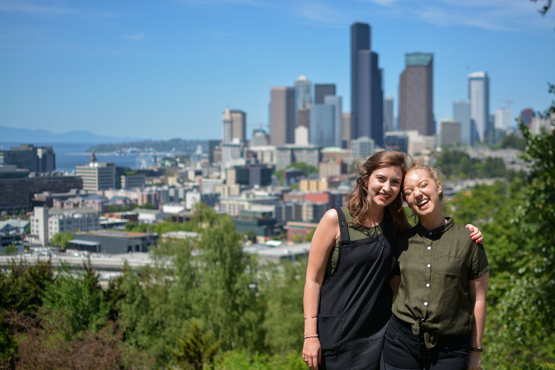 Two people smiling, posing with a city skyline in the background. Bright sunny day.